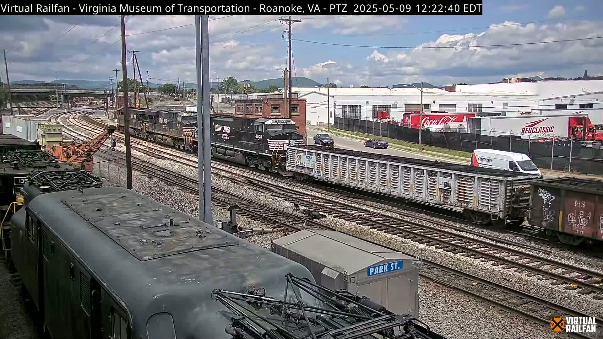 A partly cloudy day reveals a railway scene with multiple freight trains, including one with a coal car, and several industrial buildings in the background.