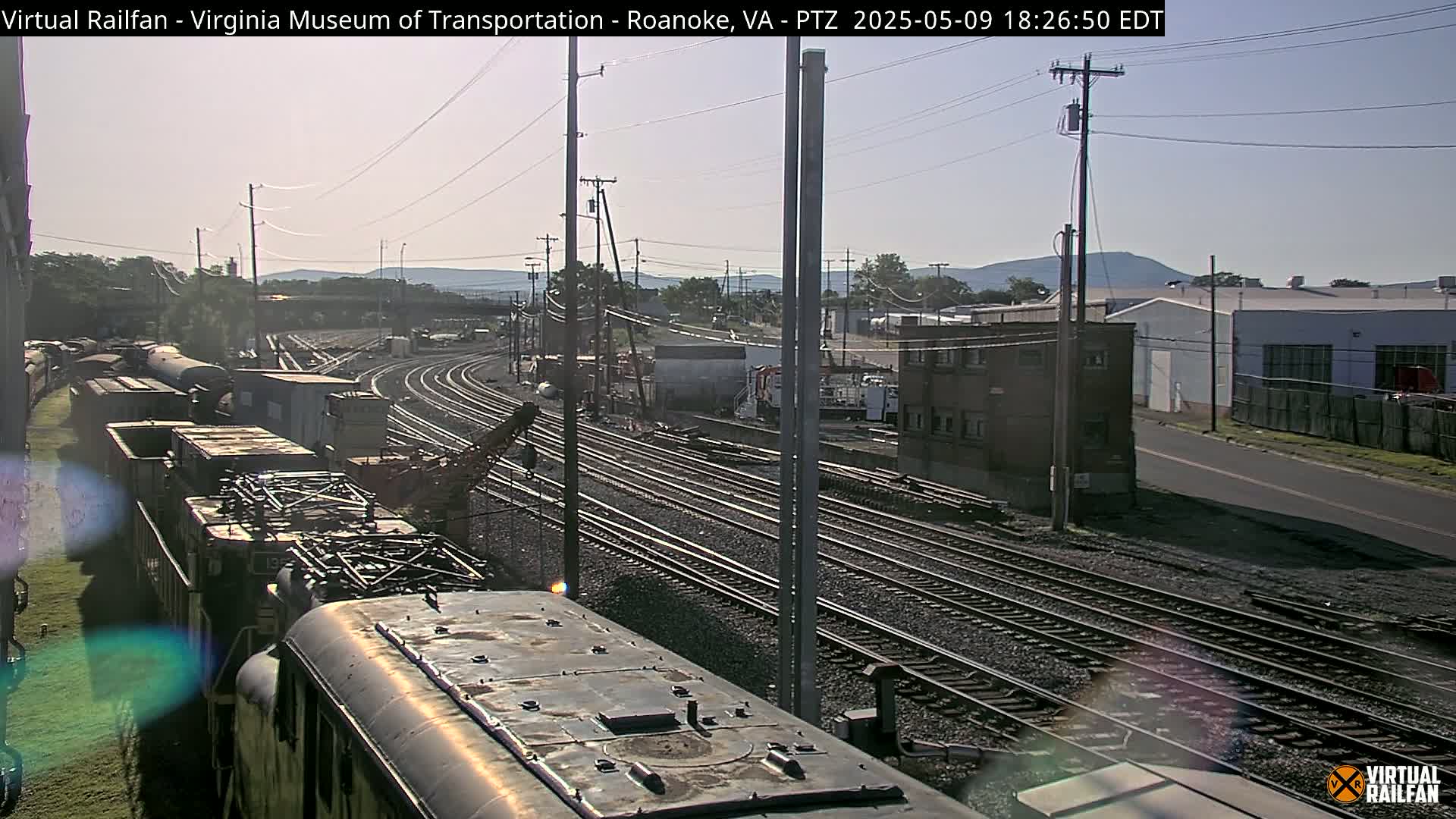 A sunny view of a railway yard with multiple train tracks, freight cars, and industrial buildings in the background.