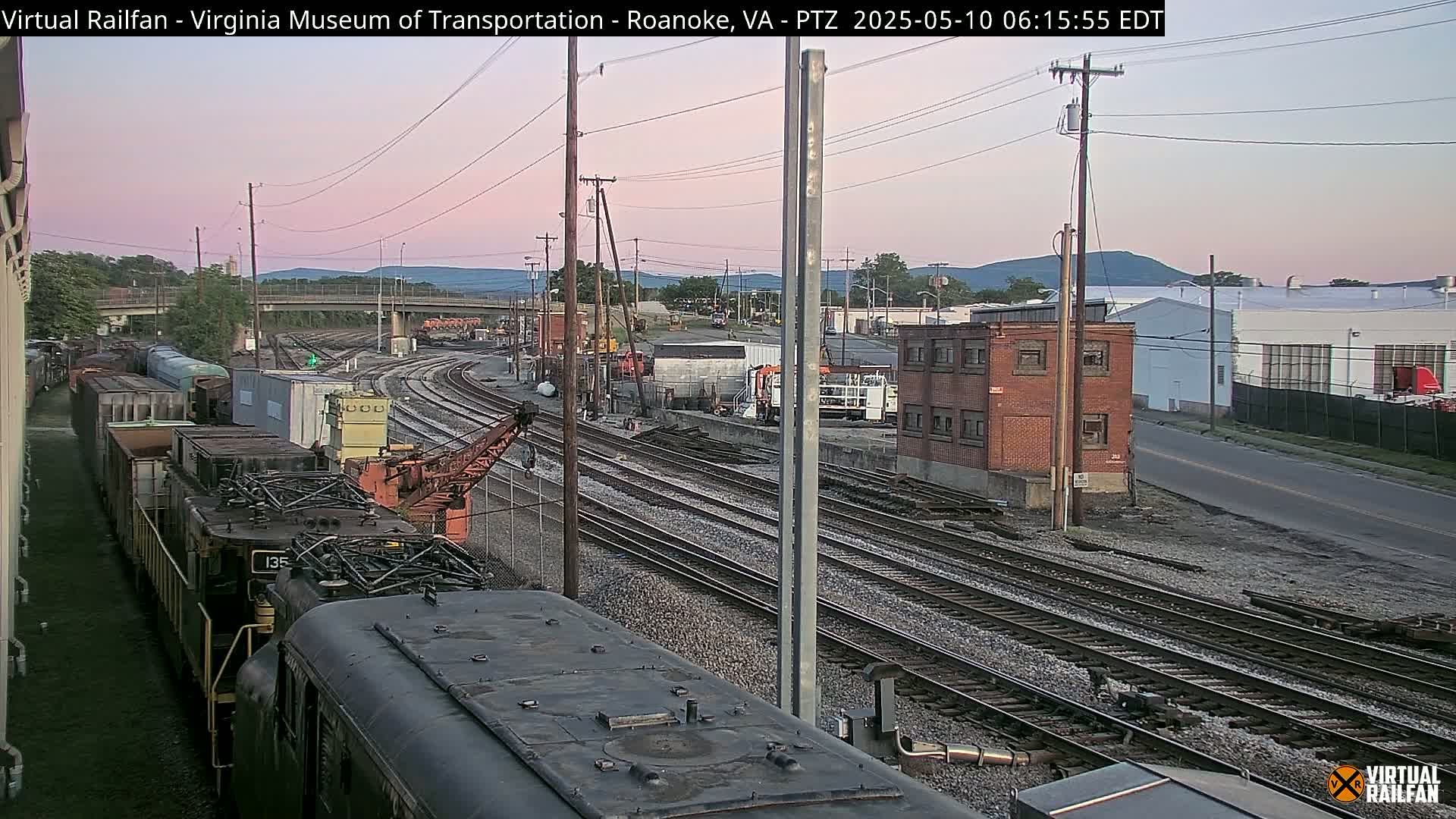 A train yard at dawn, with multiple train cars and tracks visible, under a pale pink sky.
