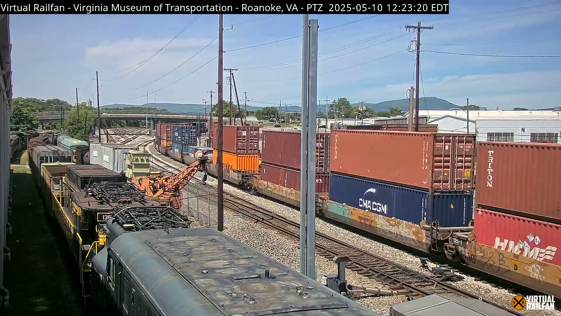 A sunny day reveals several trains with colorful shipping containers on railway tracks, with one train partially obscured in the foreground.