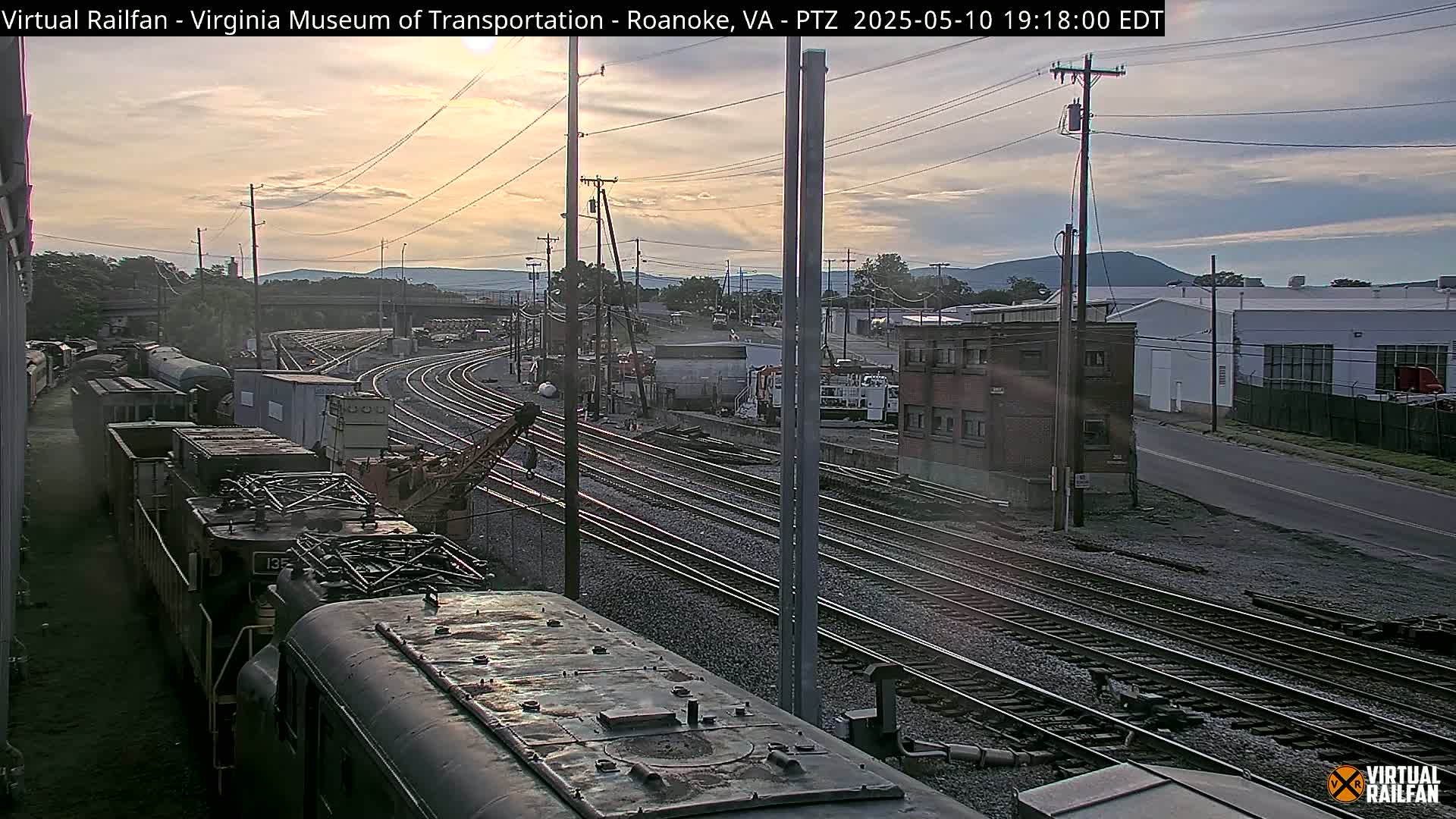 A train yard at sunset shows multiple train cars and tracks, with a few buildings and distant mountains visible in the background.