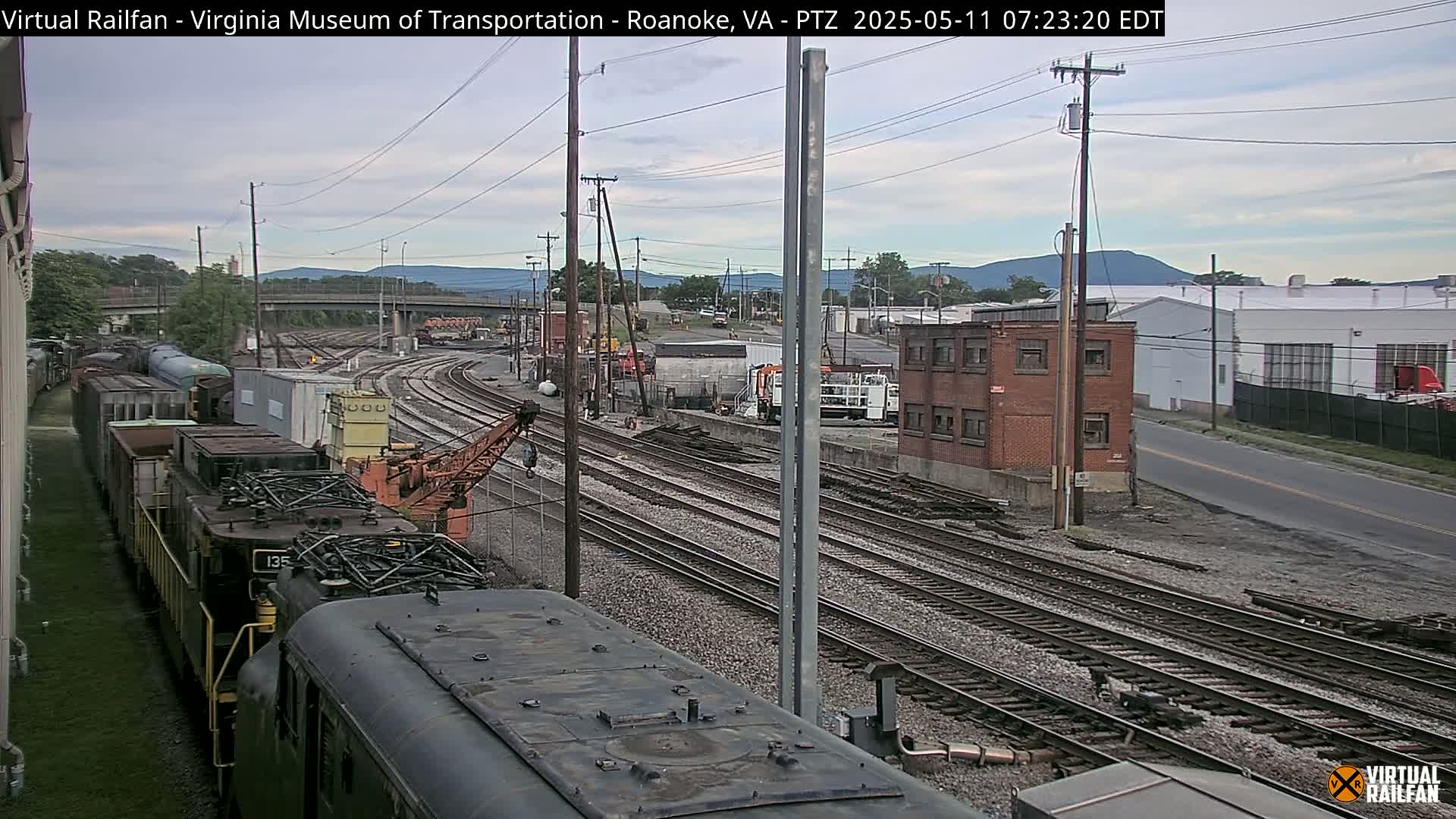 A train yard with several locomotives and freight cars, under a partly cloudy sky, is situated near industrial buildings and a highway against a backdrop of distant mountains.