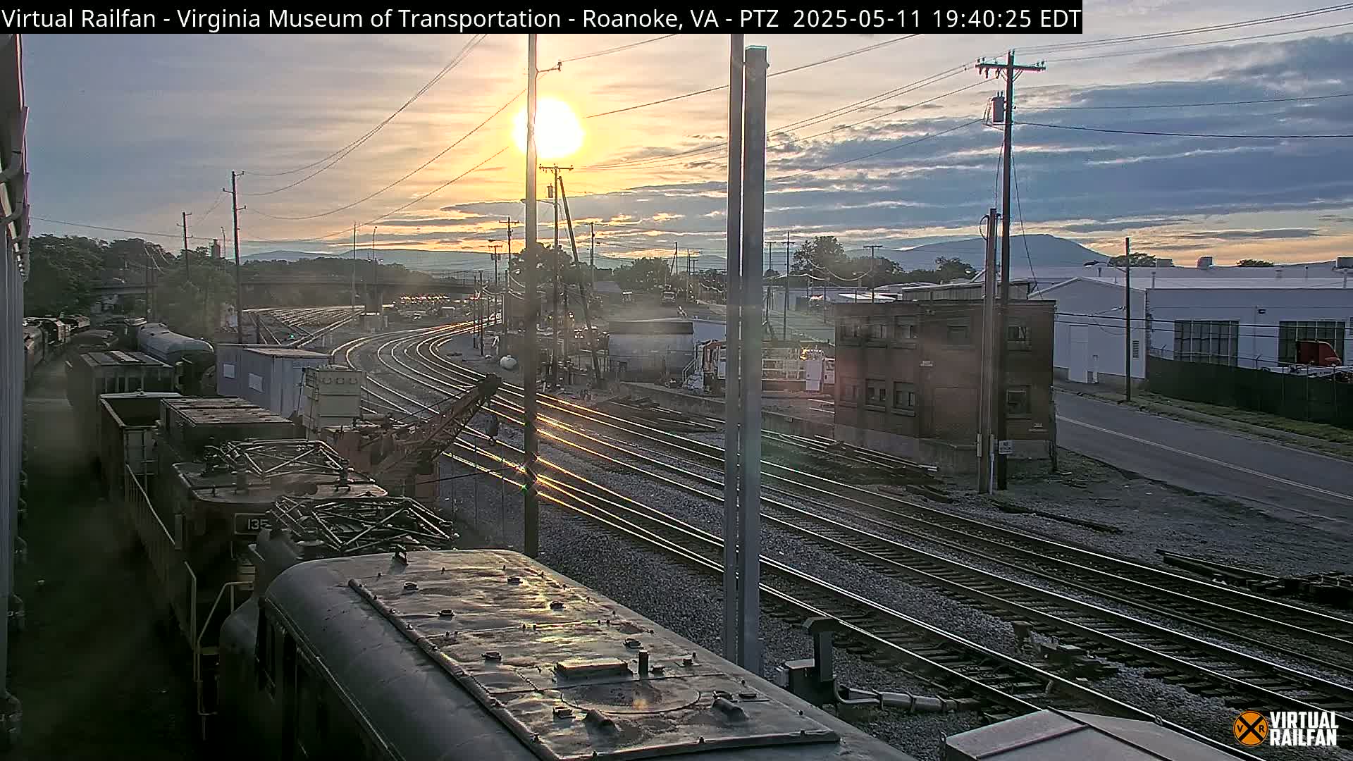 A sunset illuminates a train yard with multiple sets of railway tracks, several locomotives, and industrial buildings under a partly cloudy sky.