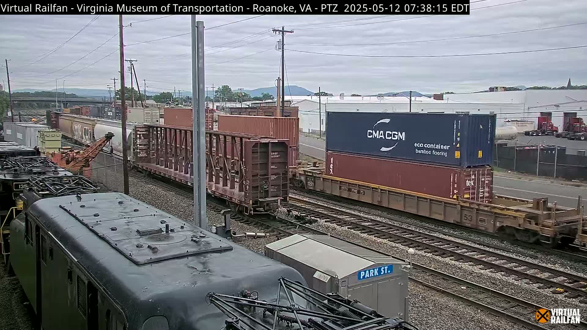 A railroad yard under an overcast sky shows several freight trains with various cargo containers, including one marked "CMA CGM," near a crane and other industrial buildings.
