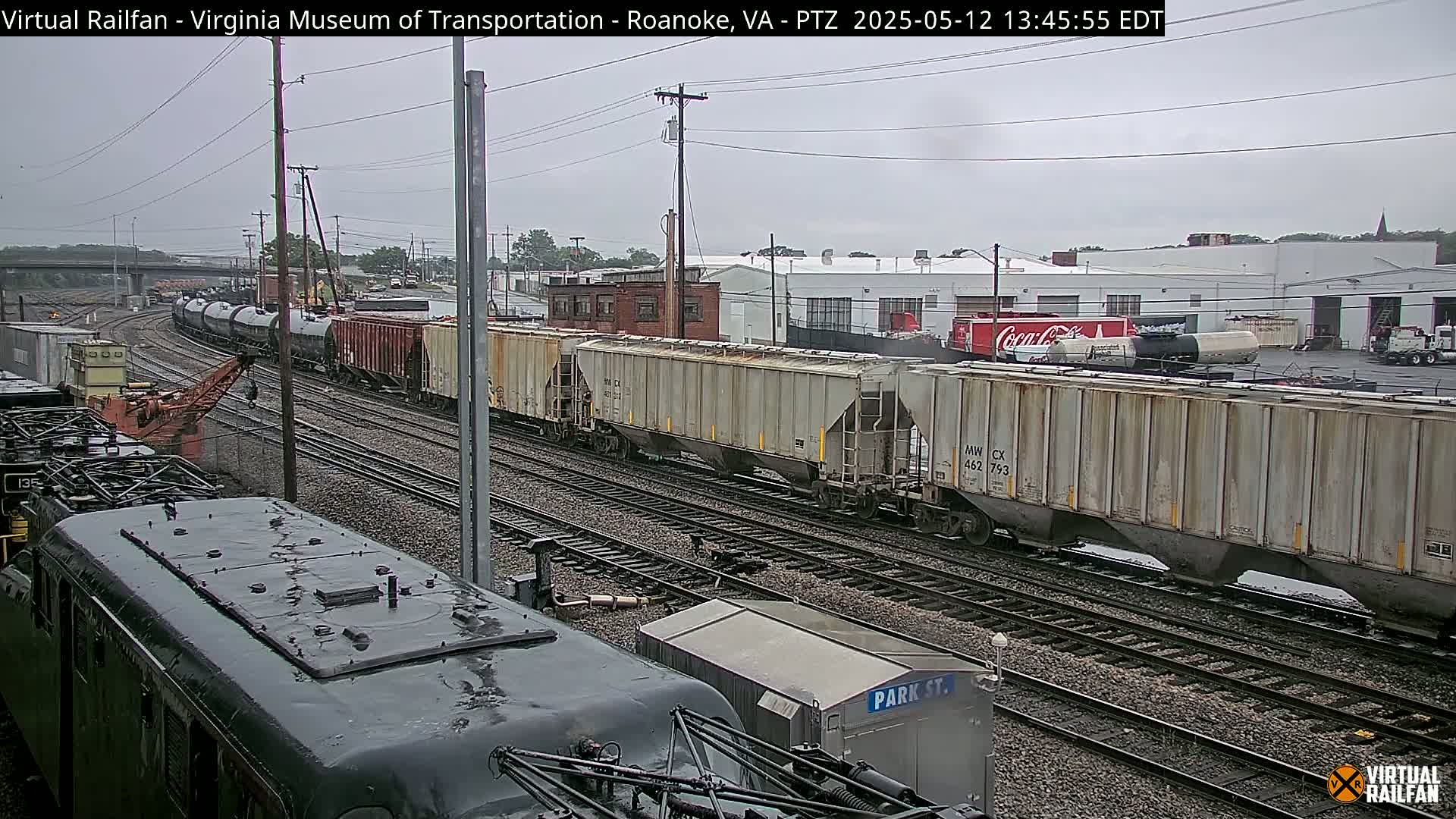A long freight train of various cars passes by industrial buildings under an overcast sky.