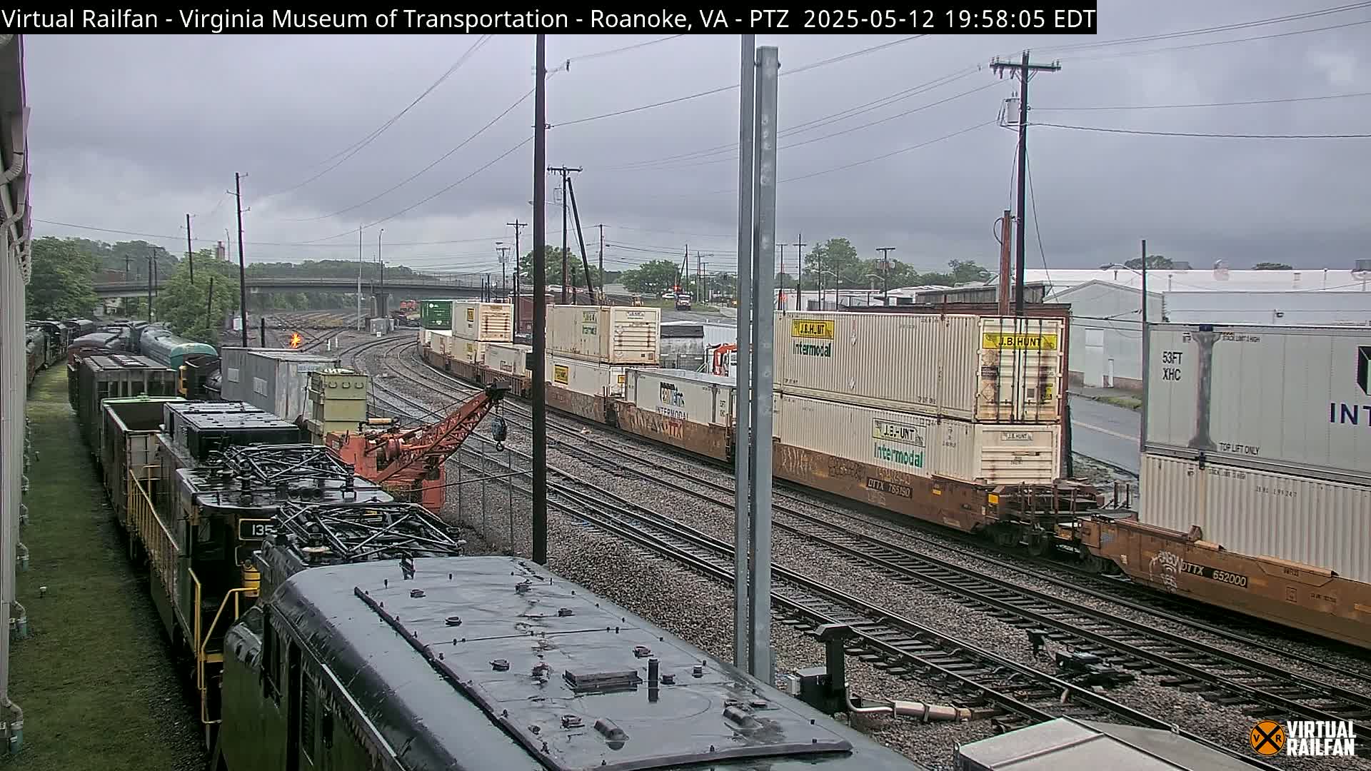 A rainy day view of a railroad yard with multiple freight trains and containers, one of which is being worked on by a crane.