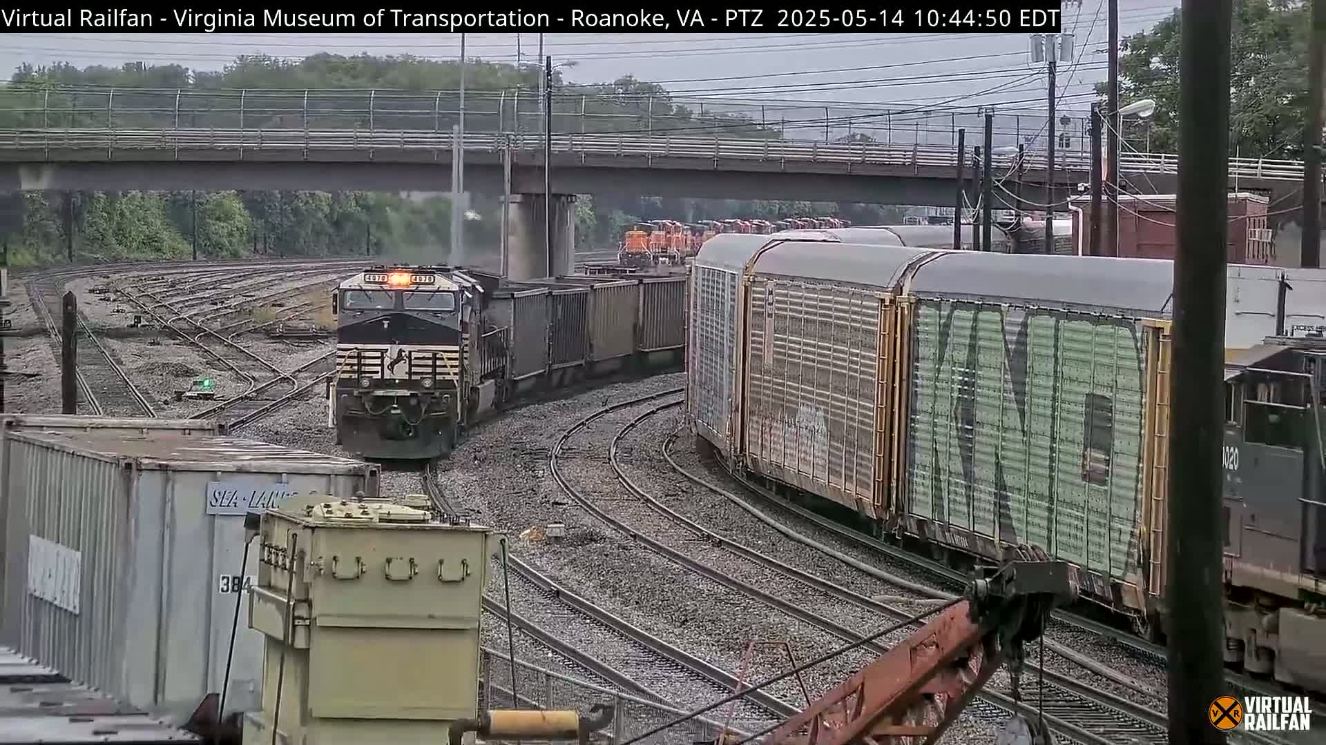 A long freight train, led by a black locomotive, curves around a track under an overpass in overcast conditions.