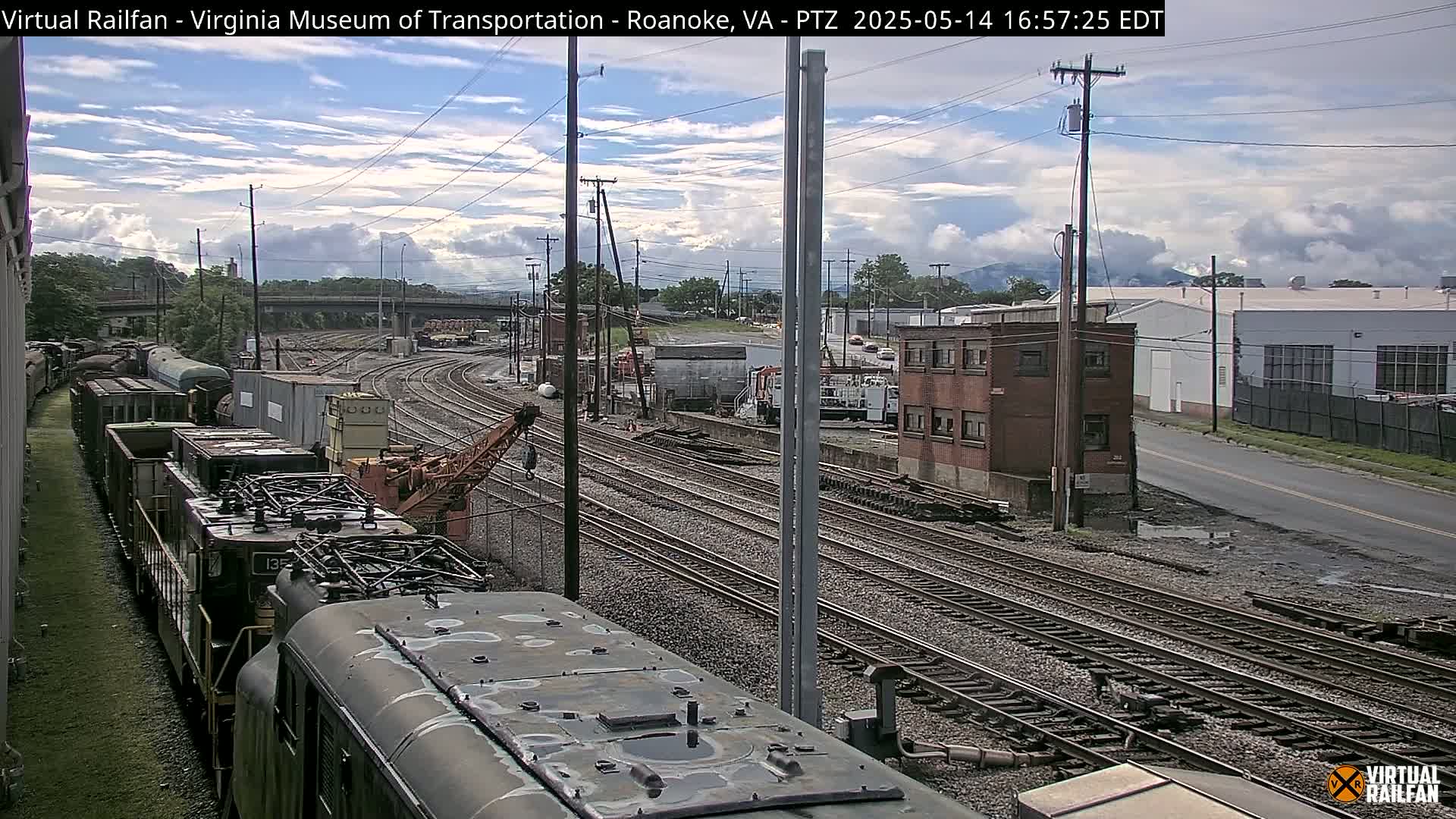 A partly cloudy day reveals a railway yard with multiple train cars and tracks, a crane, and industrial buildings in the background.