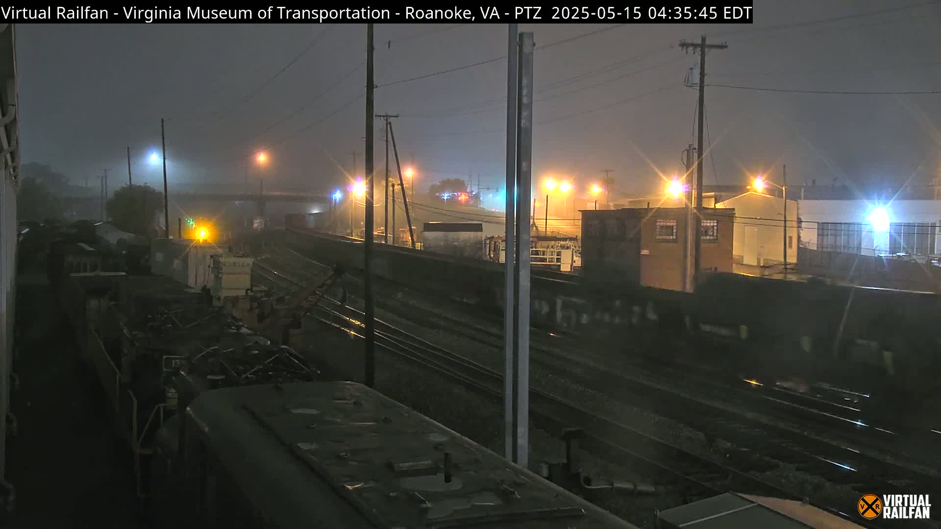 A nighttime view of railroad tracks and industrial buildings under foggy conditions, with several trains visible.