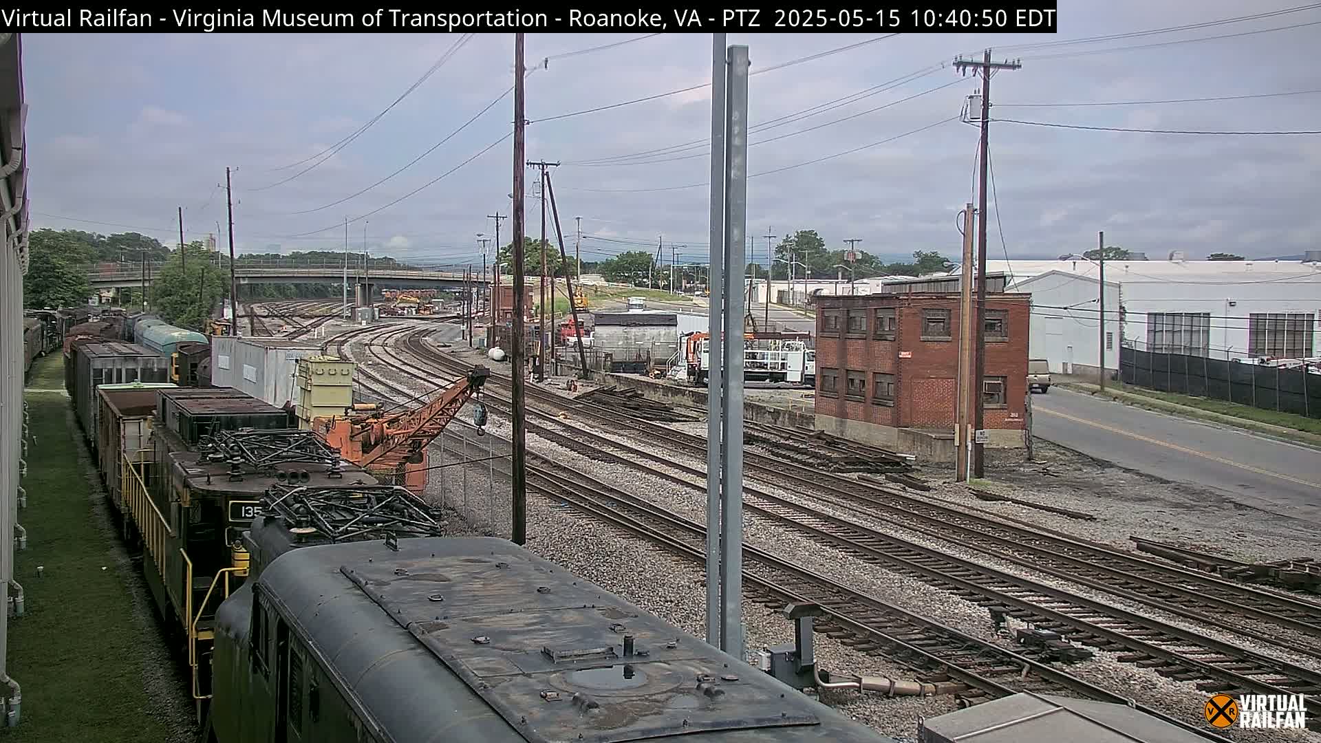 A train yard with multiple sets of railway tracks, a partially visible train, and several industrial buildings under an overcast sky.