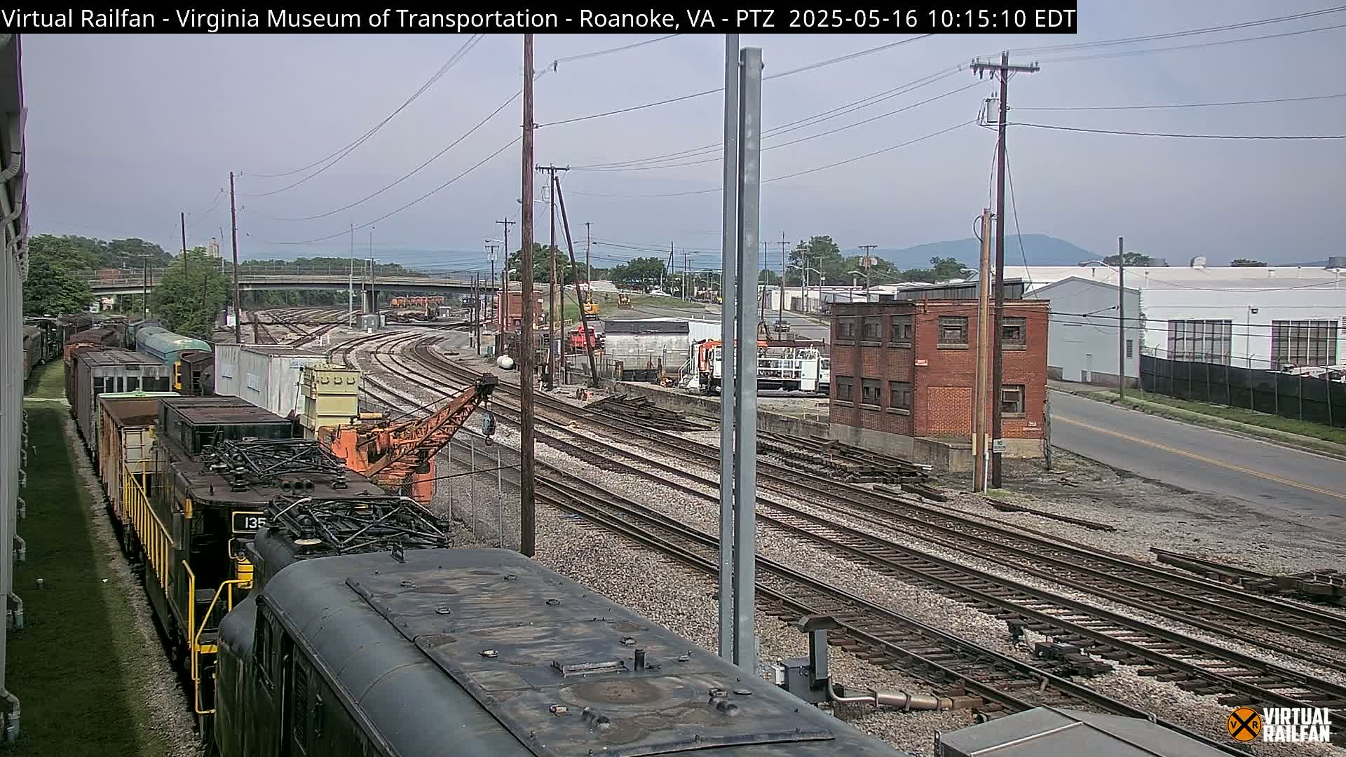 A mostly sunny day reveals a railway yard with multiple train cars, a crane, and industrial buildings, seen from atop a train.