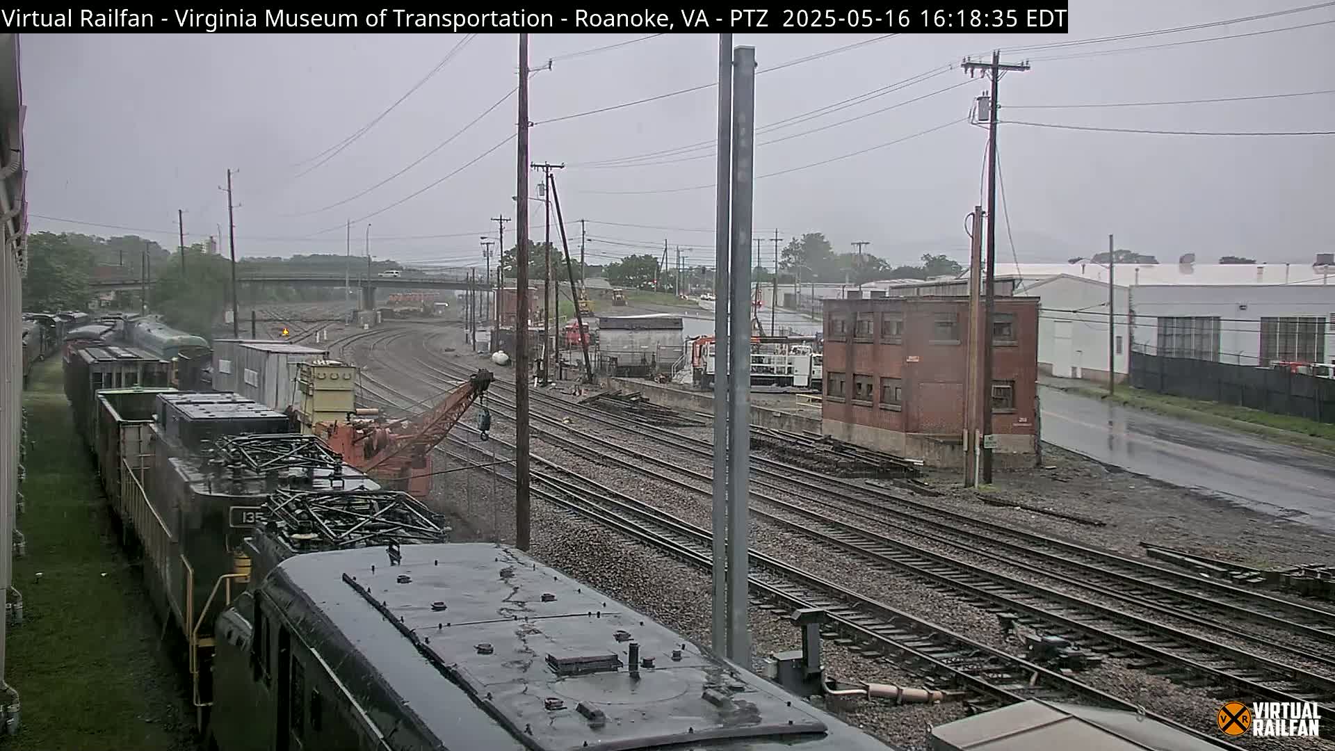 A rainy view of a railroad yard with several locomotives and train cars, a crane, and industrial buildings in the background.