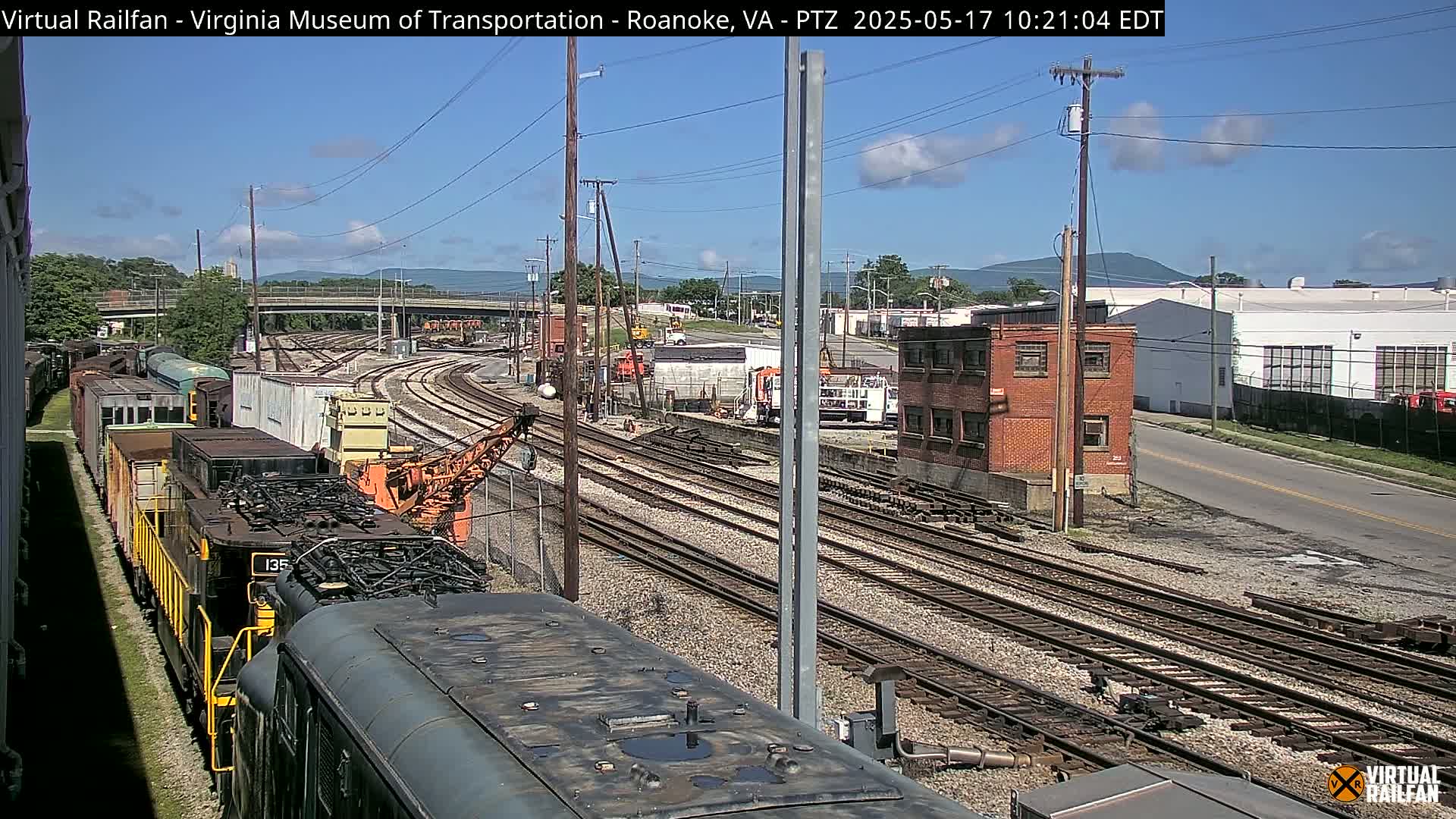A sunny day overlooking a railway yard with multiple train cars and tracks, alongside industrial buildings and a road, with mountains in the background.