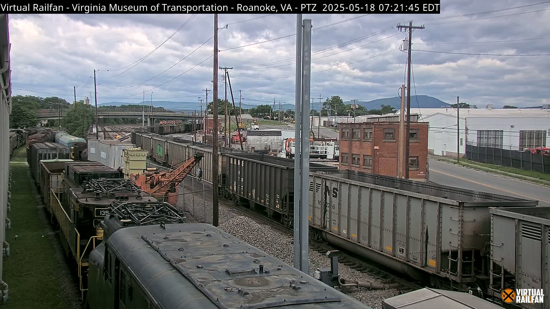 A long freight train is parked on a rail line under a cloudy sky, with industrial buildings and distant mountains visible in the background.