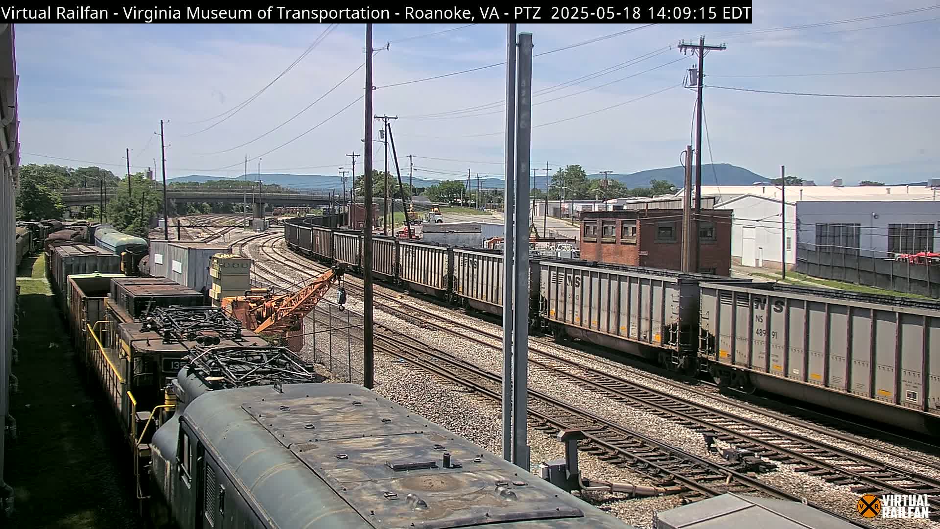 Under a sunny sky, several freight trains are visible on a network of railroad tracks, with industrial buildings and distant mountains in the background.
