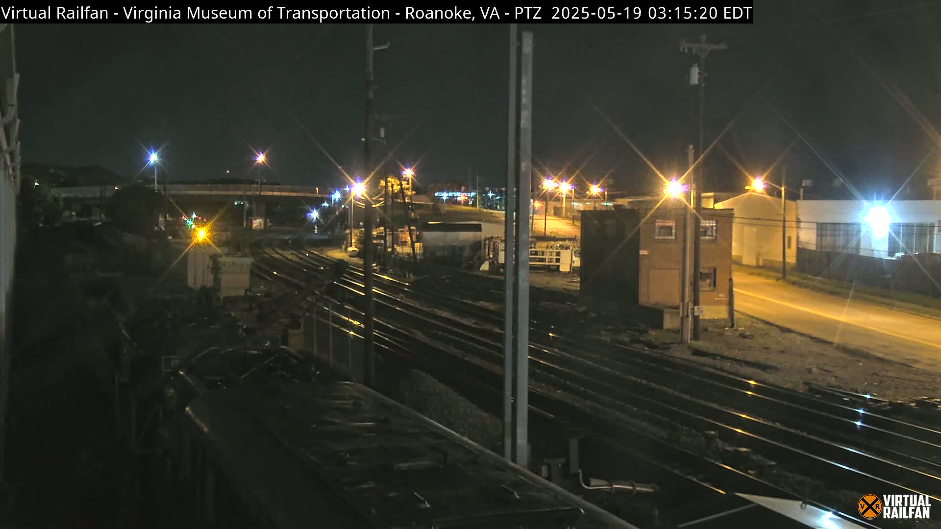 At night, a view from a train shows multiple sets of train tracks extending into the distance, passing alongside some buildings and under a street, lit by streetlights.