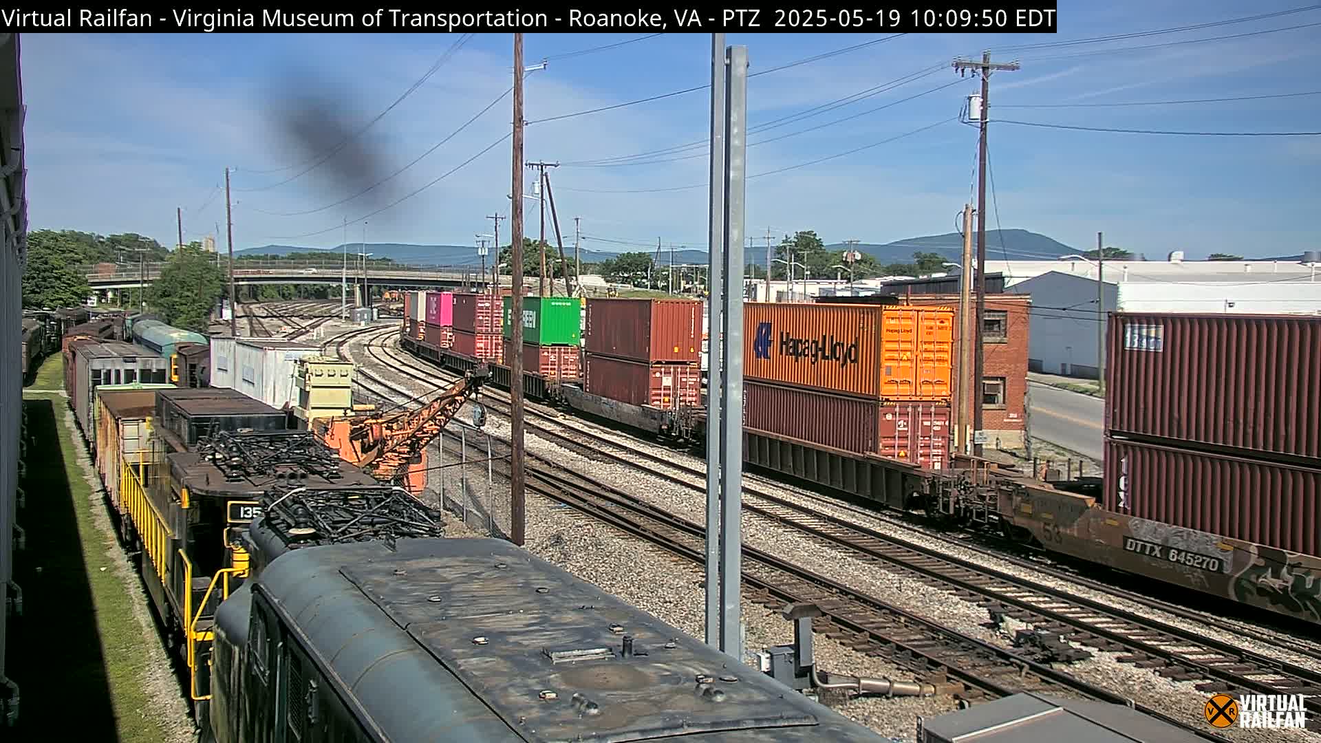 A sunny day reveals a train yard with several freight trains and colorful shipping containers, some of which are stacked two high, on multiple parallel train tracks.