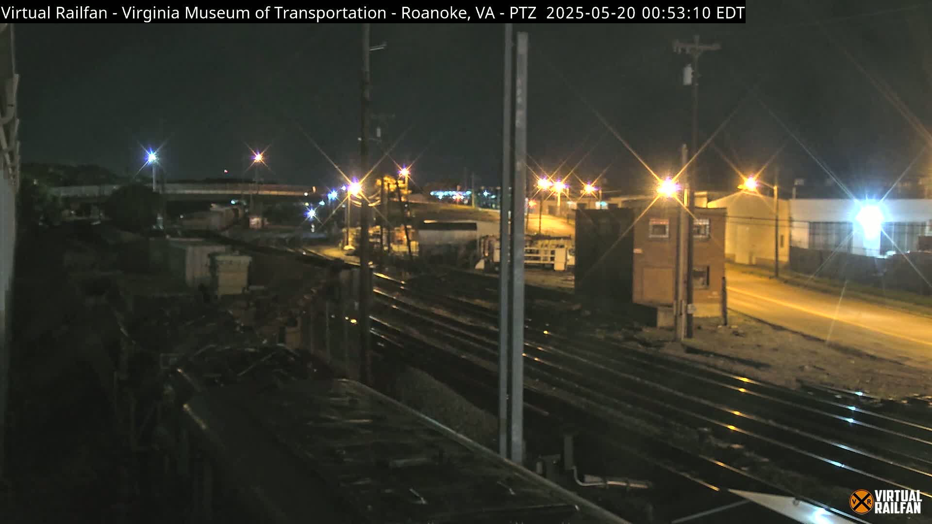A nighttime view of railroad tracks and a street, illuminated by streetlights, with industrial buildings and a train partially visible in the foreground.