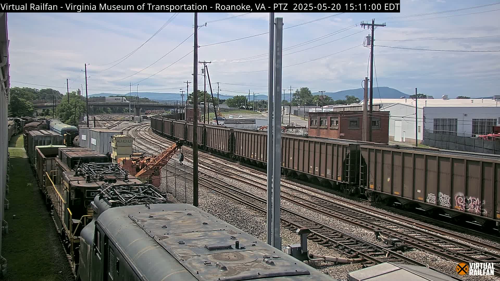 A mostly sunny day reveals a railway yard with multiple freight trains, including locomotives and hopper cars, situated amidst industrial buildings and a distant mountain range.