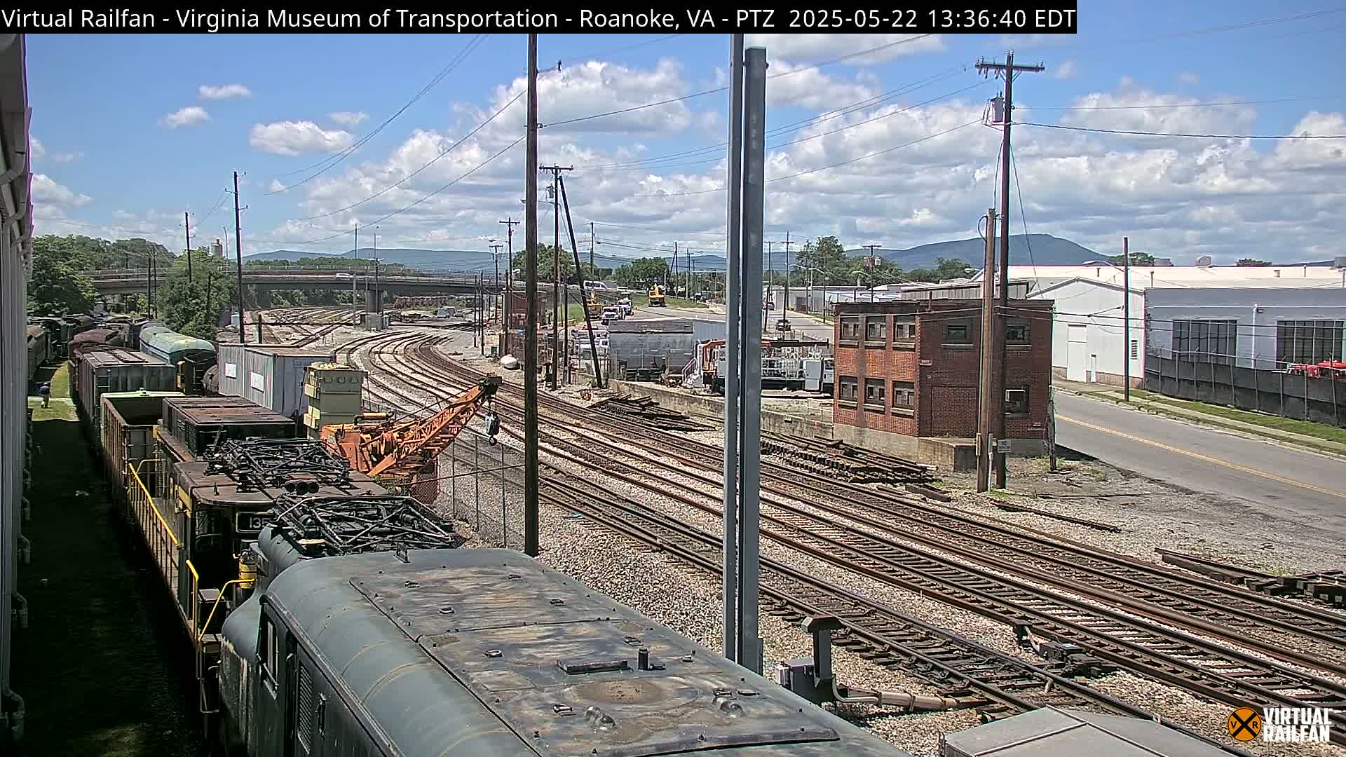 A partly cloudy day reveals a railroad yard with multiple train cars and tracks, a crane, and several buildings in the background.