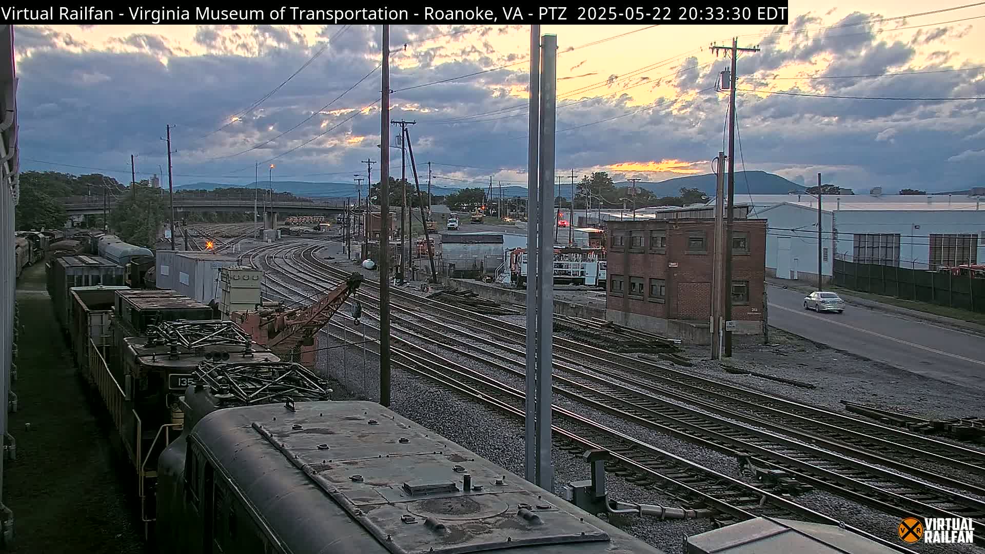 A twilight view of a railroad yard with several train cars, a crane, and a brick building, under a partly cloudy sky.
