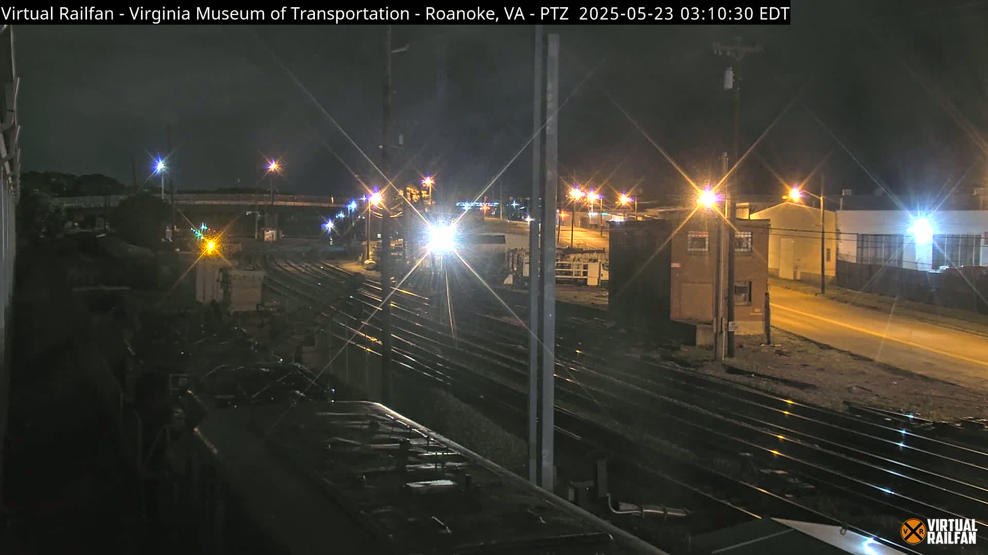 A nighttime view of a train yard with multiple sets of tracks, illuminated by streetlights and the headlights of a train in the distance.