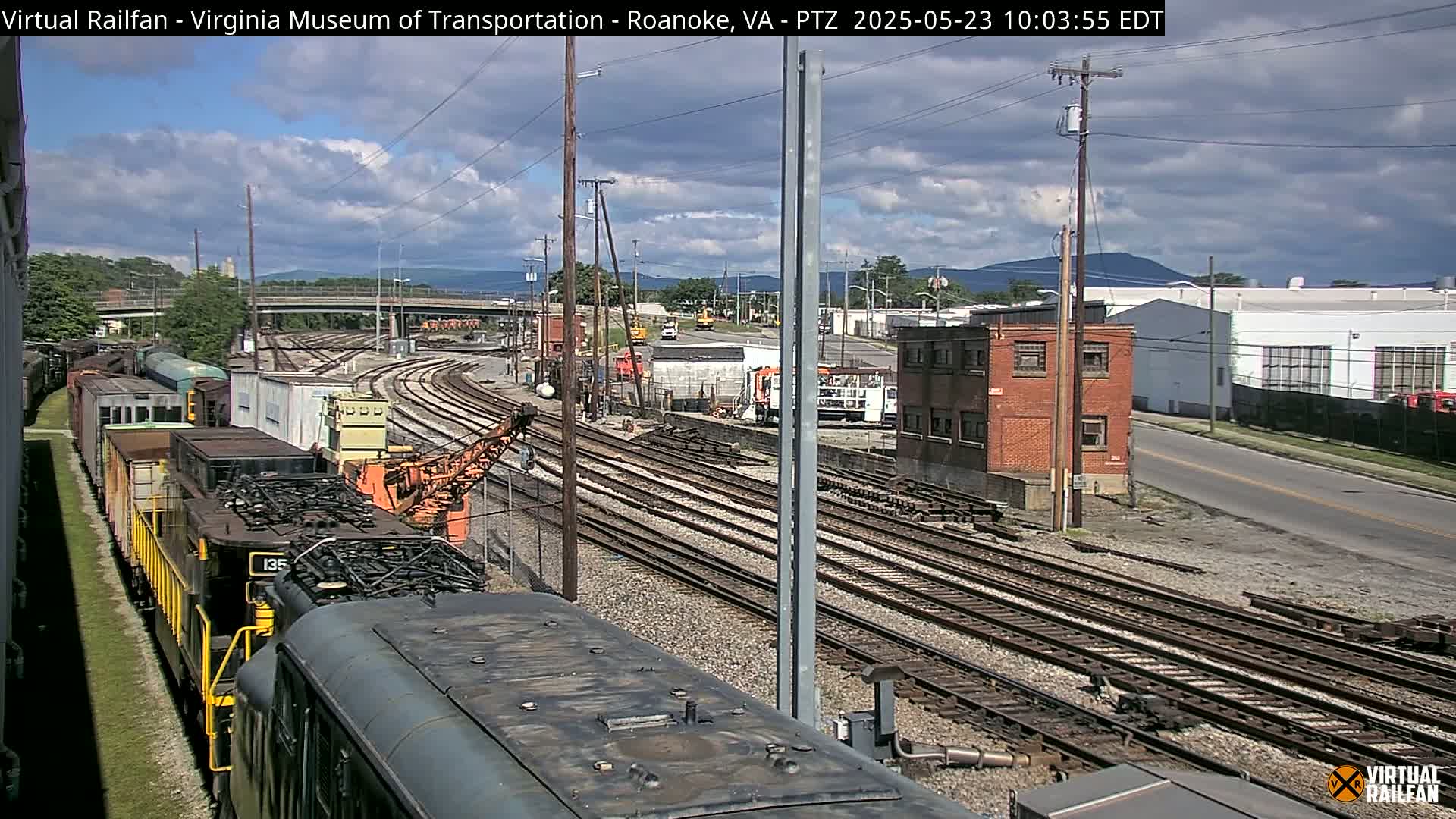 A partly cloudy day reveals a railroad yard with multiple train tracks, a locomotive, and industrial buildings under a partly cloudy sky.