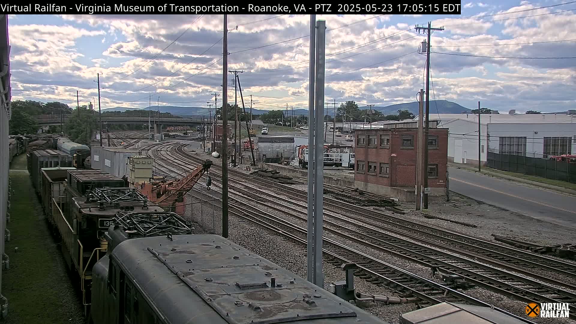 A partly cloudy day shows a rail yard with several train cars and locomotives, a crane, and industrial buildings, all under a partly cloudy sky.