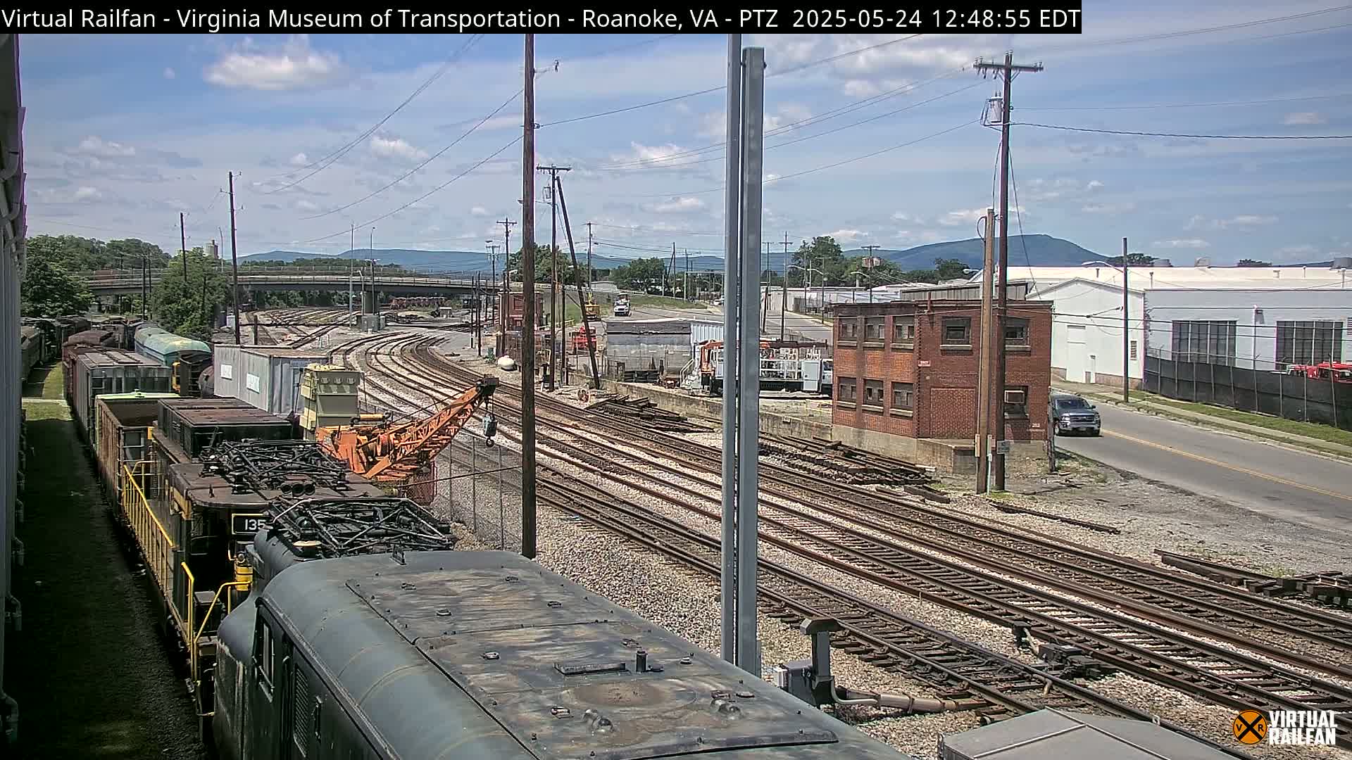 A sunny day shows a railroad yard with multiple train tracks, railway cars, and a crane, next to a street and industrial buildings with distant mountains.