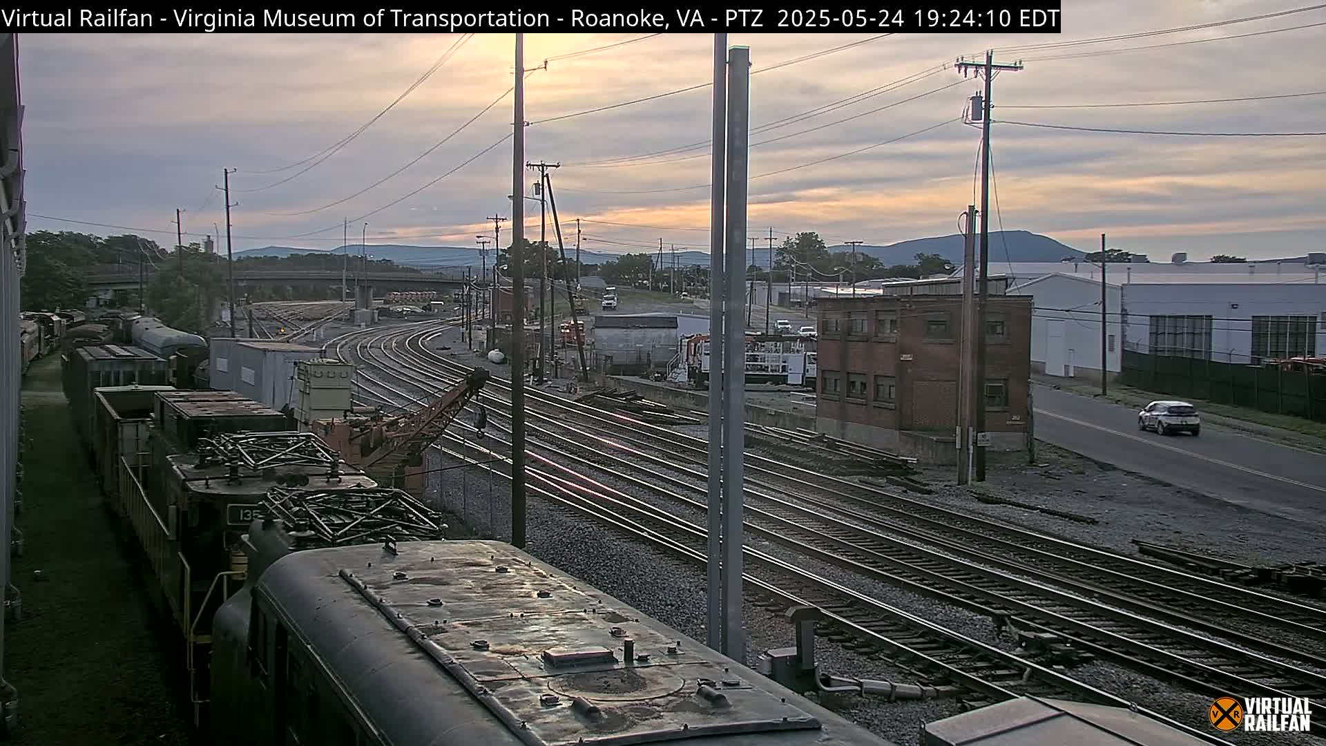 A twilight view of a train yard with multiple railway tracks, several train cars, and a single car driving on a nearby road under a partly cloudy sky.