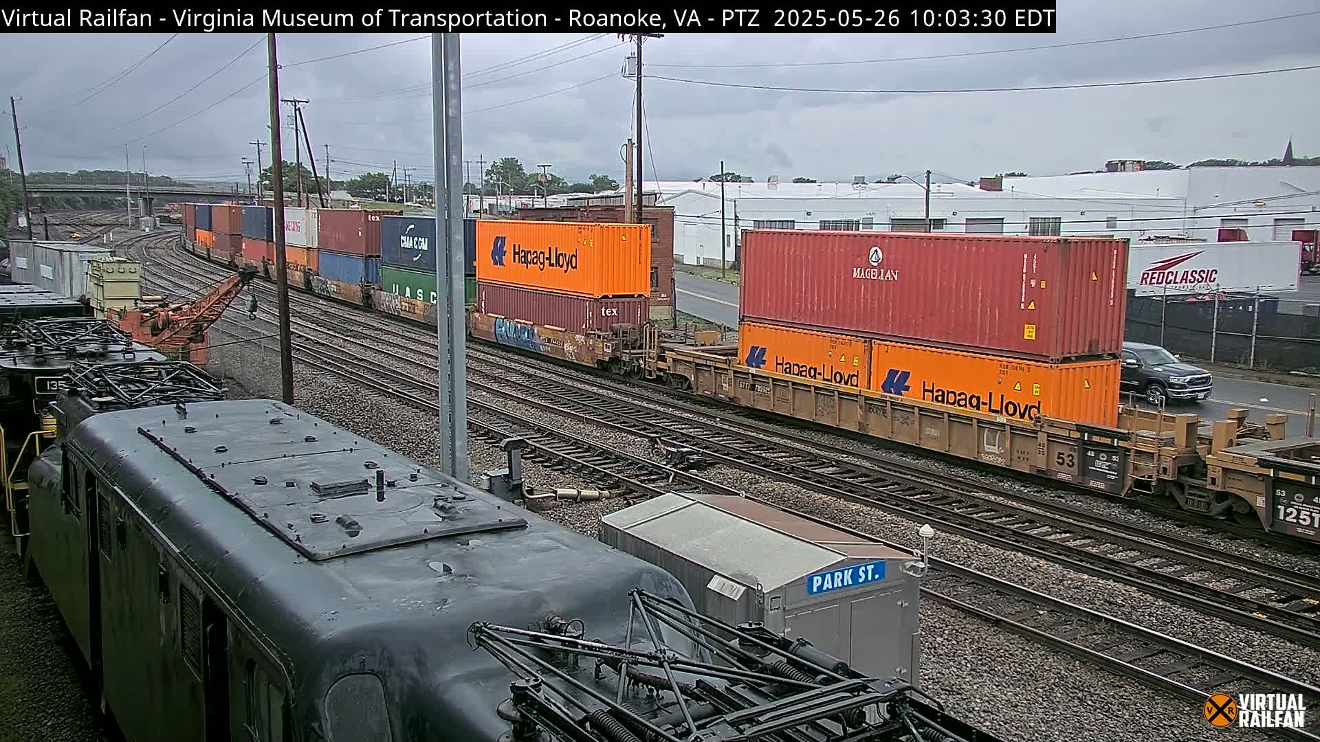A cloudy day reveals multiple freight trains with various shipping containers on parallel railroad tracks, one of which is partially obscured by a dark-colored locomotive.