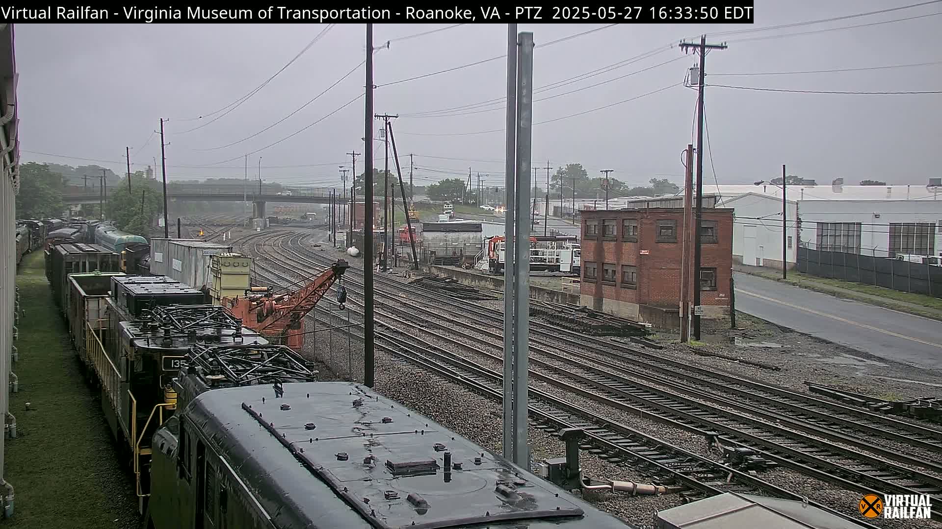 A rain-soaked railway yard with multiple train cars and tracks, a crane, and industrial buildings in the background.