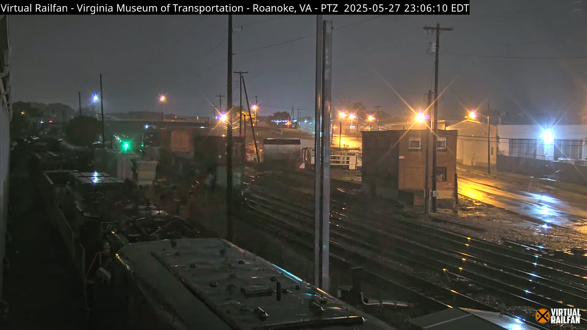 A nighttime view of rain-slicked railroad tracks and surrounding buildings, illuminated by streetlights and the lights of a train.
