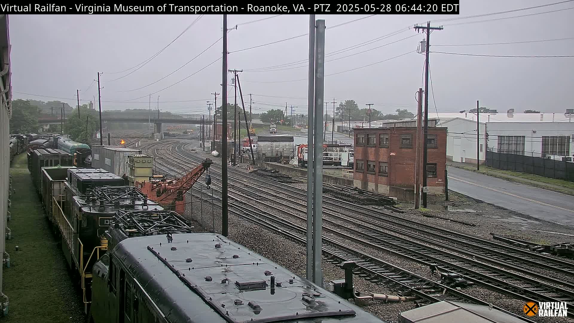 A rainy view of a train yard with multiple sets of railroad tracks, several locomotives, and industrial buildings in the background.