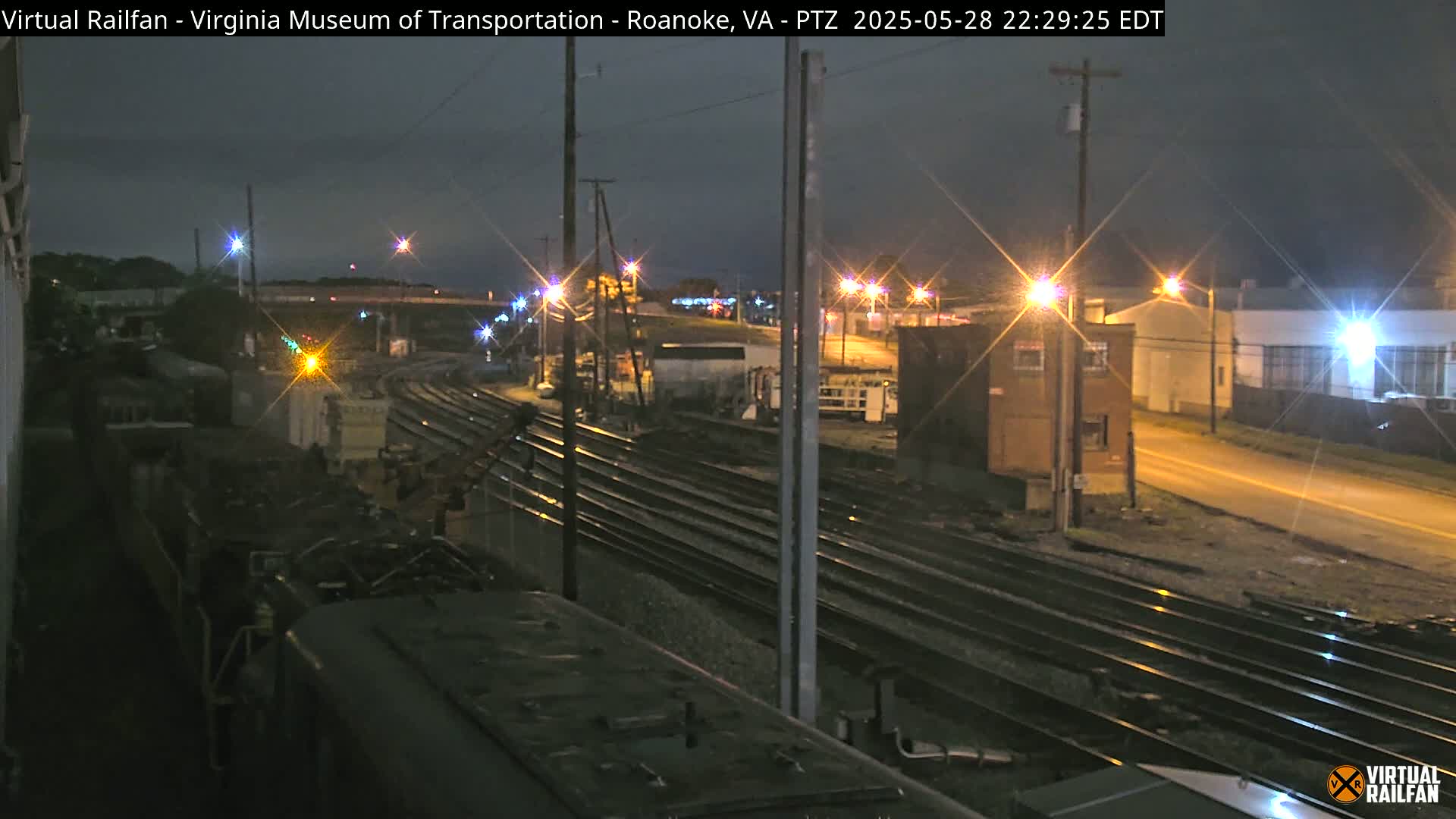 A nighttime view of a train yard with multiple sets of train tracks, illuminated by streetlights and the glow of nearby buildings.