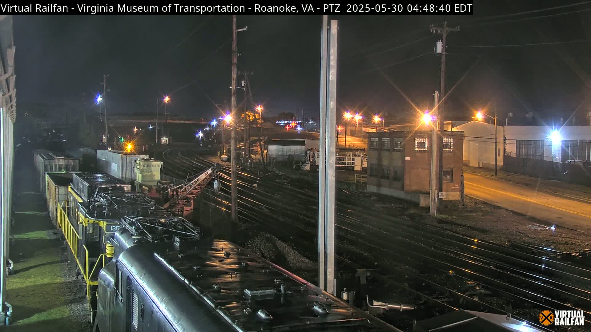 A nighttime view of a train yard with a diesel locomotive and rail cars, under streetlights and clear skies.