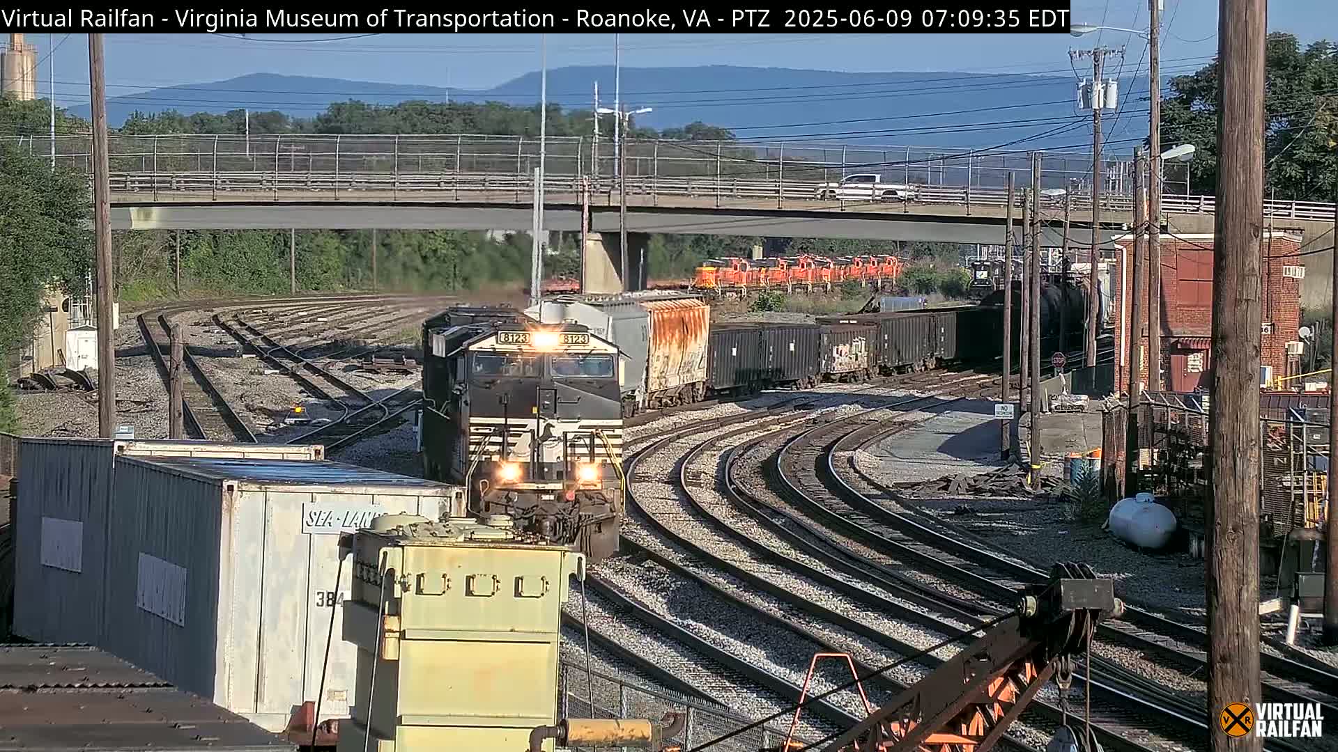 A freight train, led by a black locomotive, is navigating a curved section of railway tracks under a clear, sunny sky, with a road bridge visible in the background.