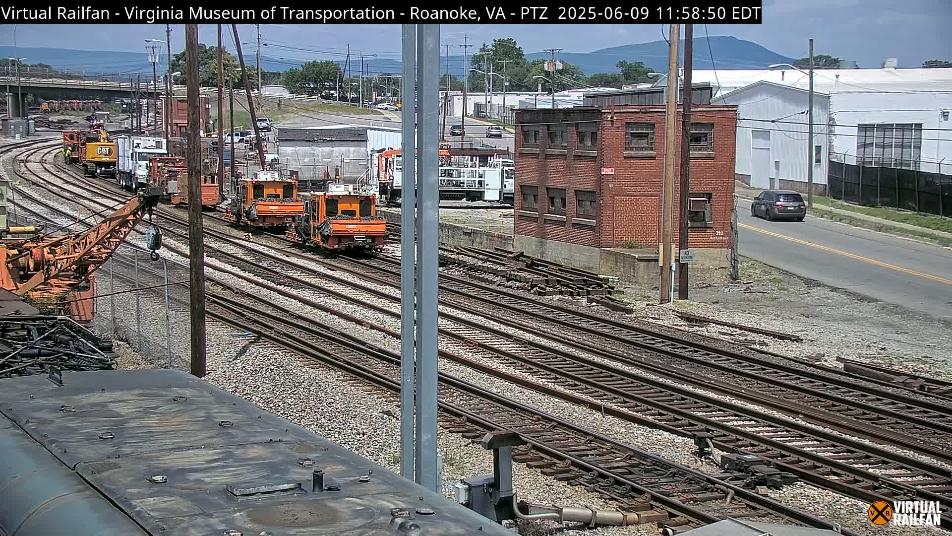 A sunny day shows several orange pieces of railway maintenance equipment parked on multiple sets of train tracks next to a brick building and a road with cars.