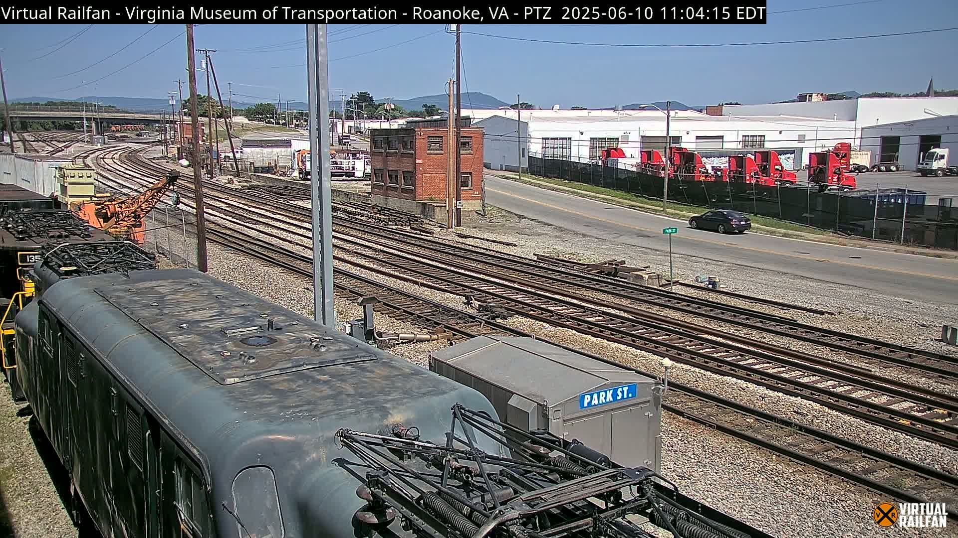 A dark-green locomotive sits at the foreground of a sunny outdoor scene of railway tracks that extend into the distance past a brick building and several red trucks.