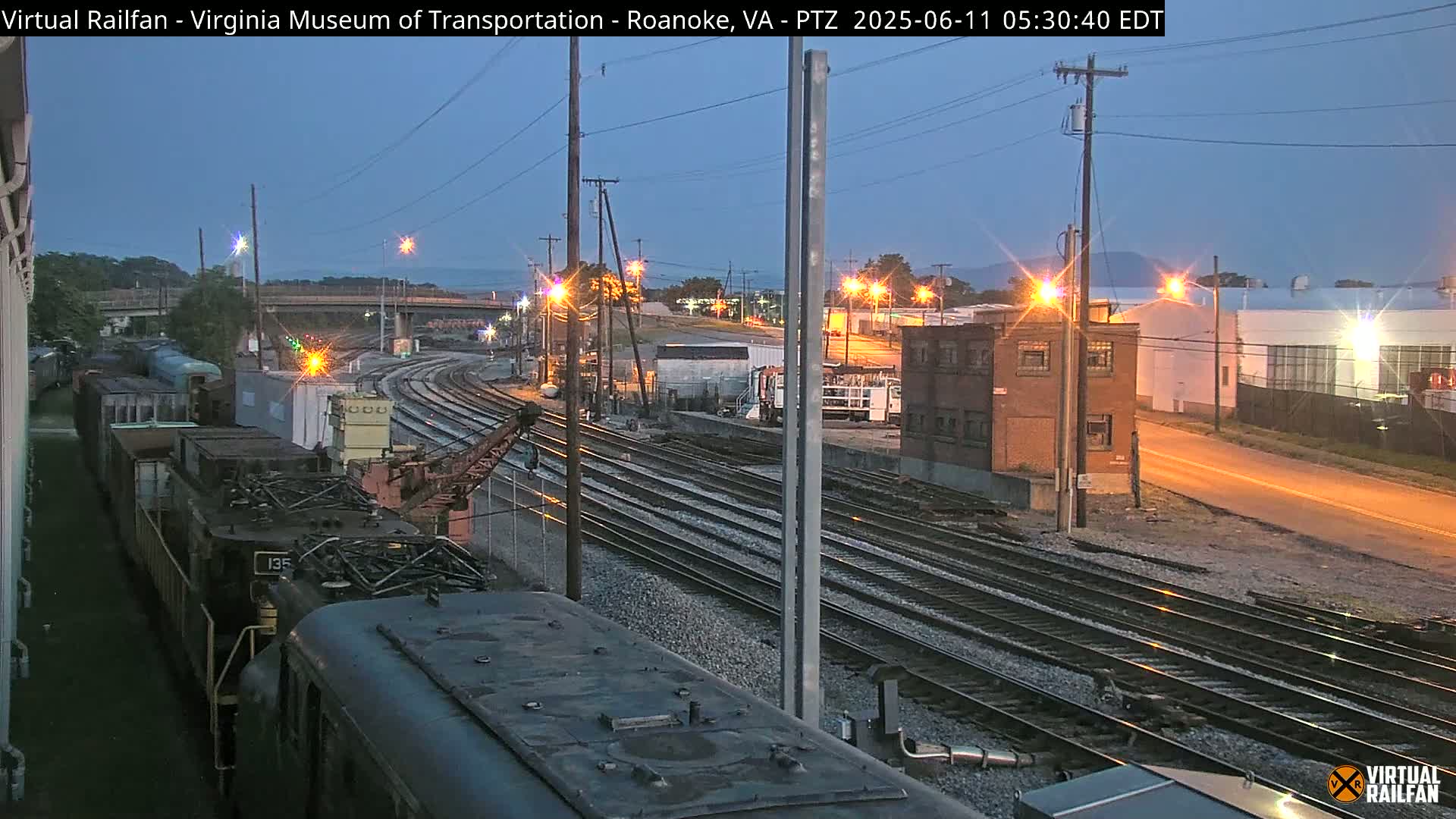 Under a twilight sky, a train sits alongside several railway tracks near industrial buildings and a street lit by streetlights.