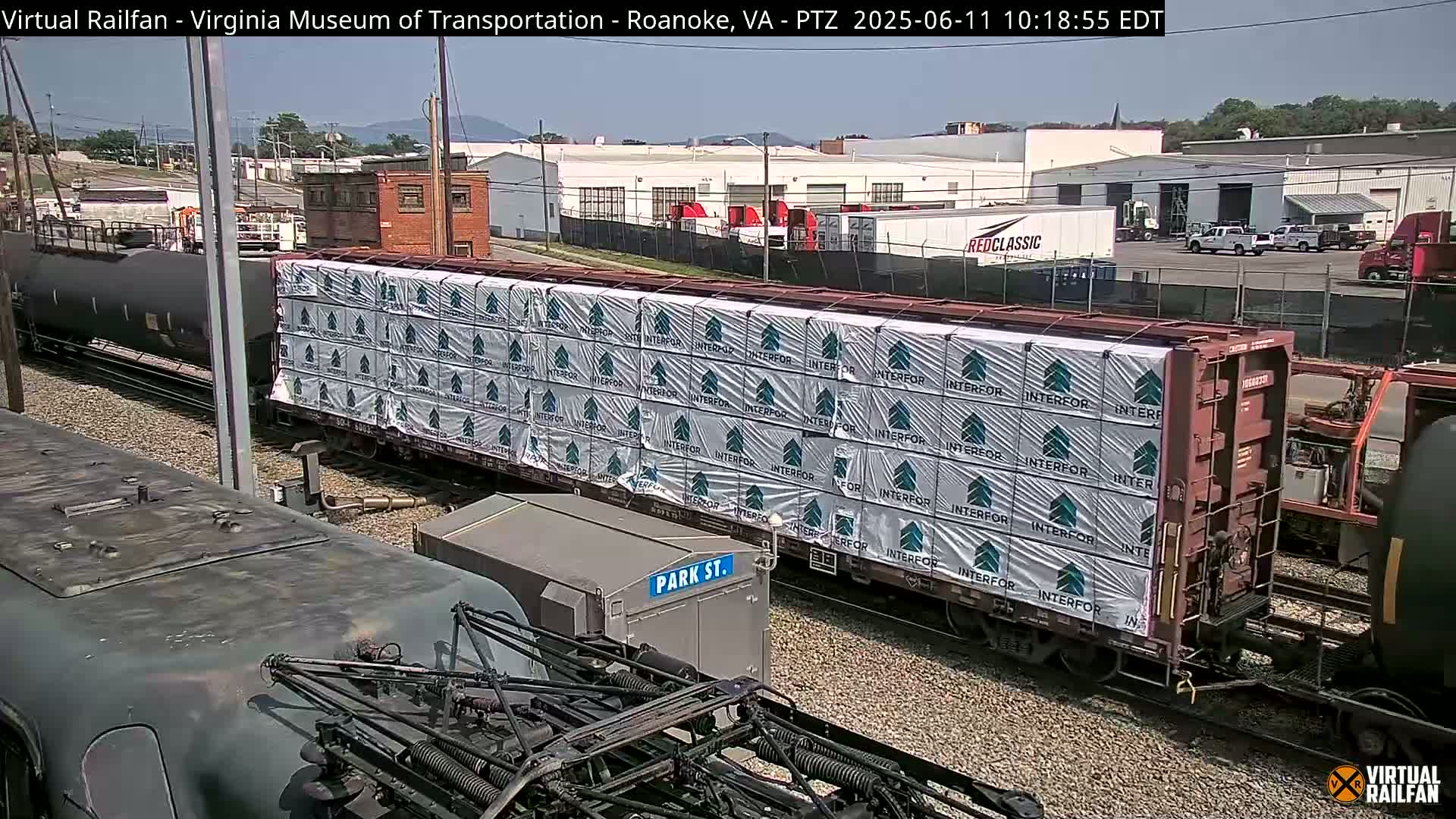 A train car loaded with bundled lumber sits on a rail line next to other train cars under a clear, sunny sky.