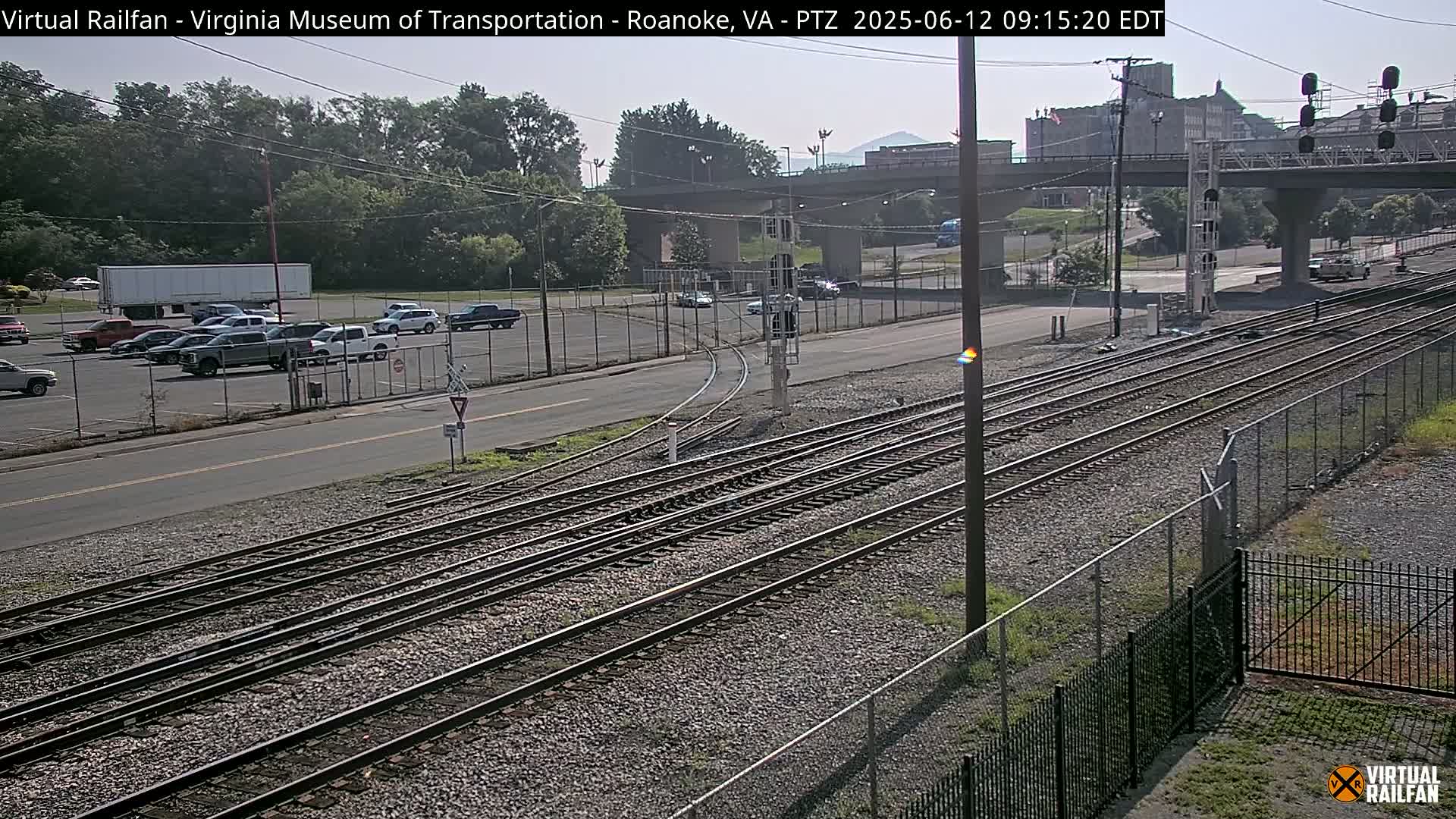 A sunny day shows a view of railroad tracks intersecting with a road, alongside a parking lot with several vehicles and a large building in the background.