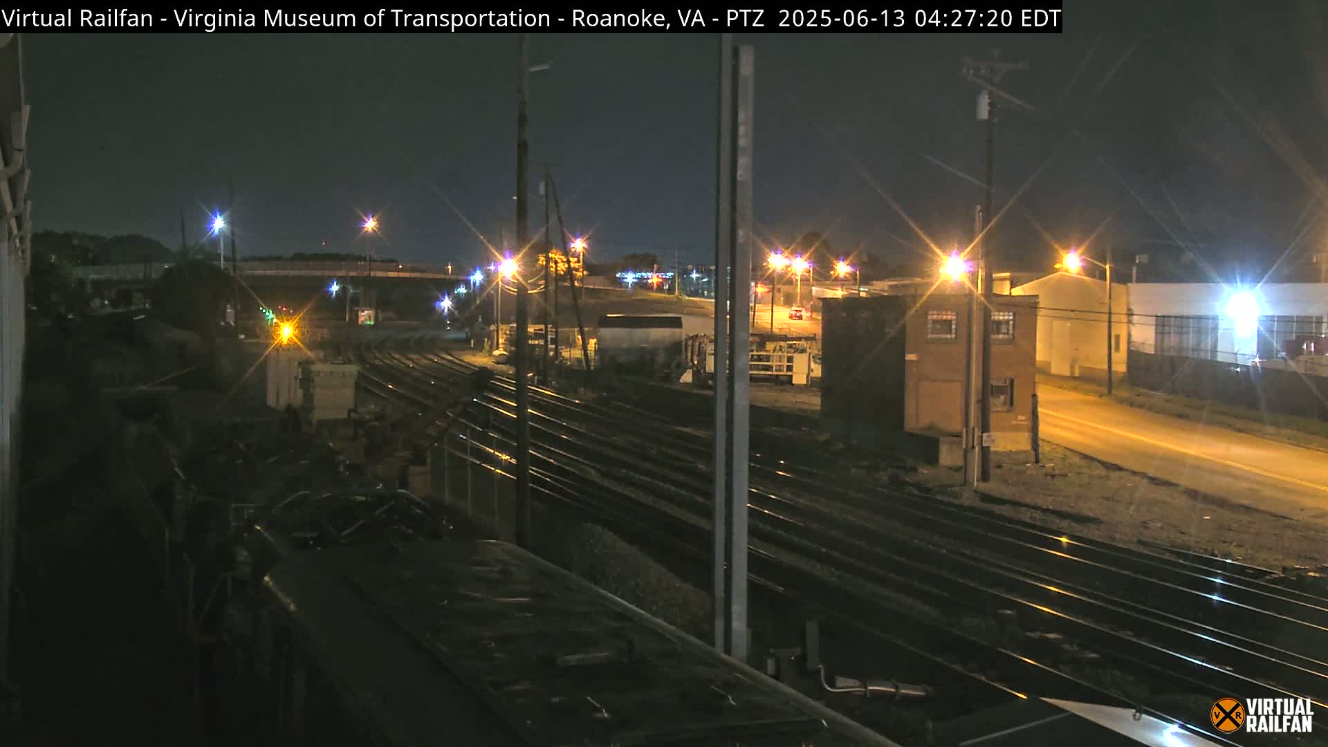 A nighttime view from atop a train car shows multiple sets of train tracks, illuminated by streetlights, alongside industrial buildings and a road.