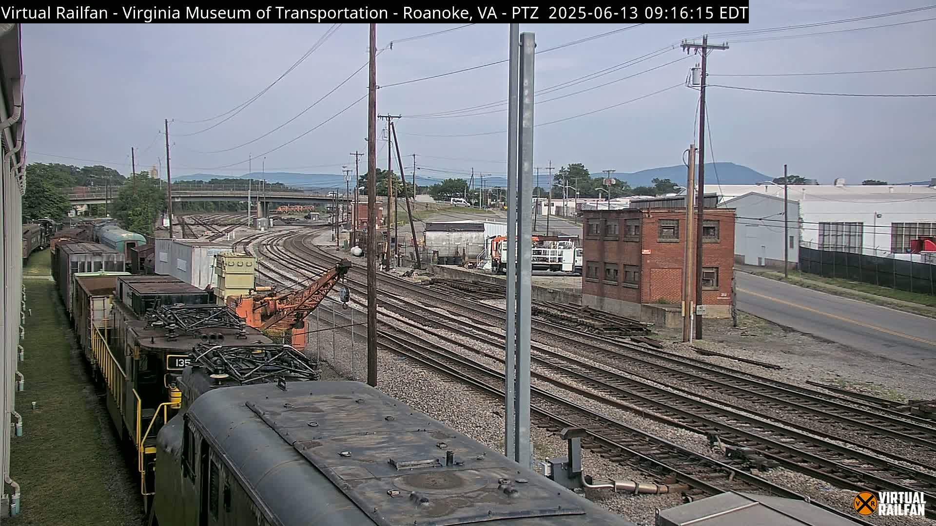 A train yard with multiple parallel railway tracks, a partially visible train, and industrial buildings under an overcast sky.