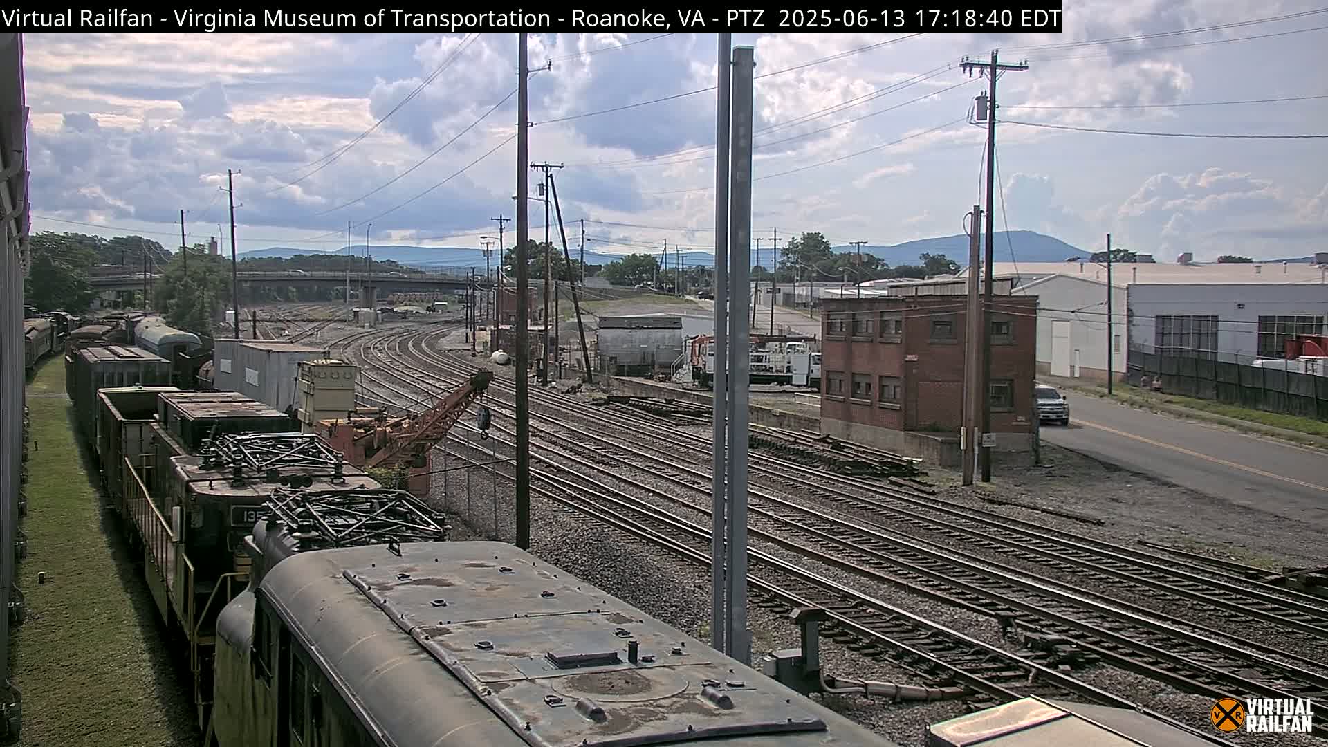 A partly cloudy day reveals a railroad yard with multiple train cars, tracks, and industrial buildings, viewed from atop a train.