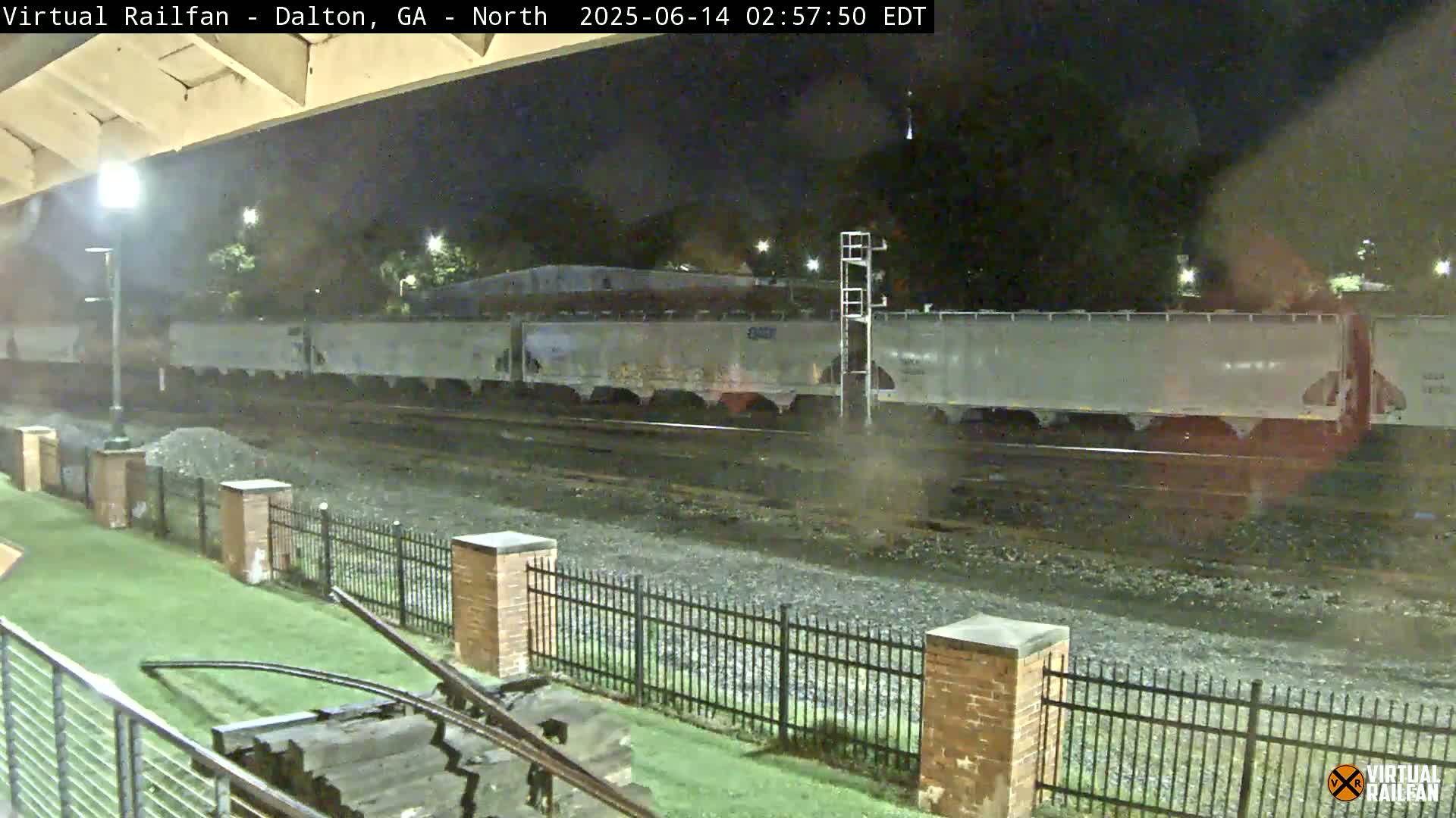 A partly cloudy day reveals a railroad yard with multiple train cars, tracks, and industrial buildings, viewed from atop a train.