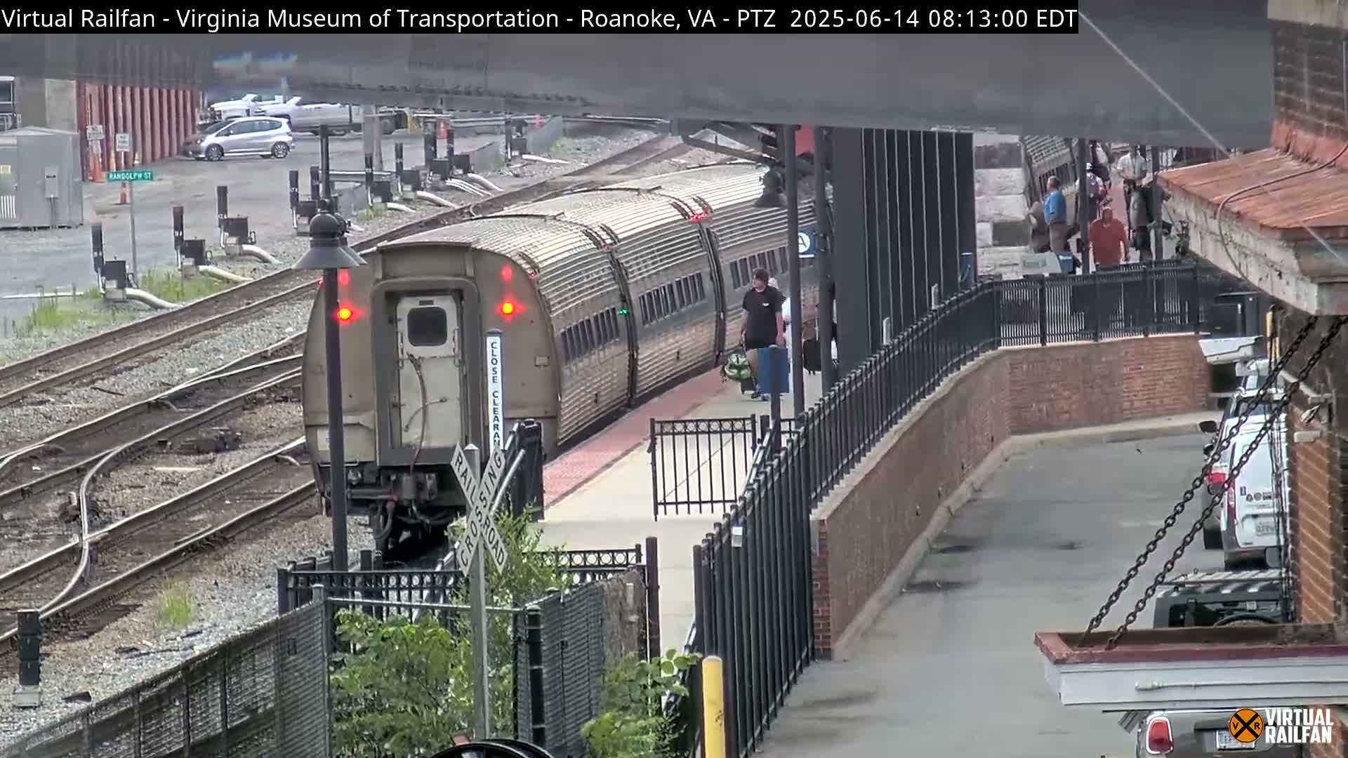 A long, light-brown passenger train is arriving at a station platform on an overcast day.