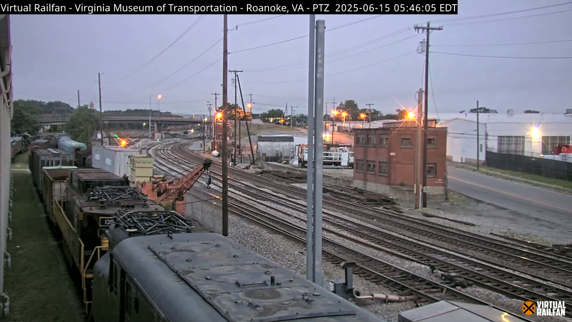 A train sits on a railway track under a twilight sky, with several other rail cars and industrial buildings in the background.