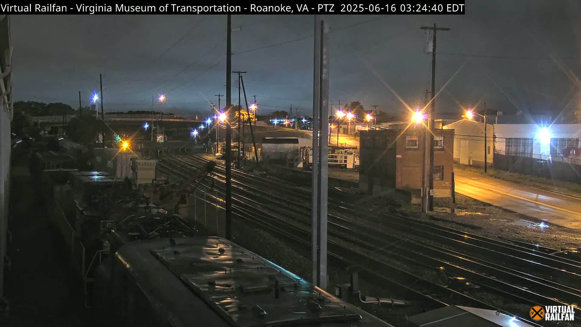 A nighttime view of railroad tracks and a train car under a dark, possibly rainy sky, with streetlights illuminating the surrounding industrial area.