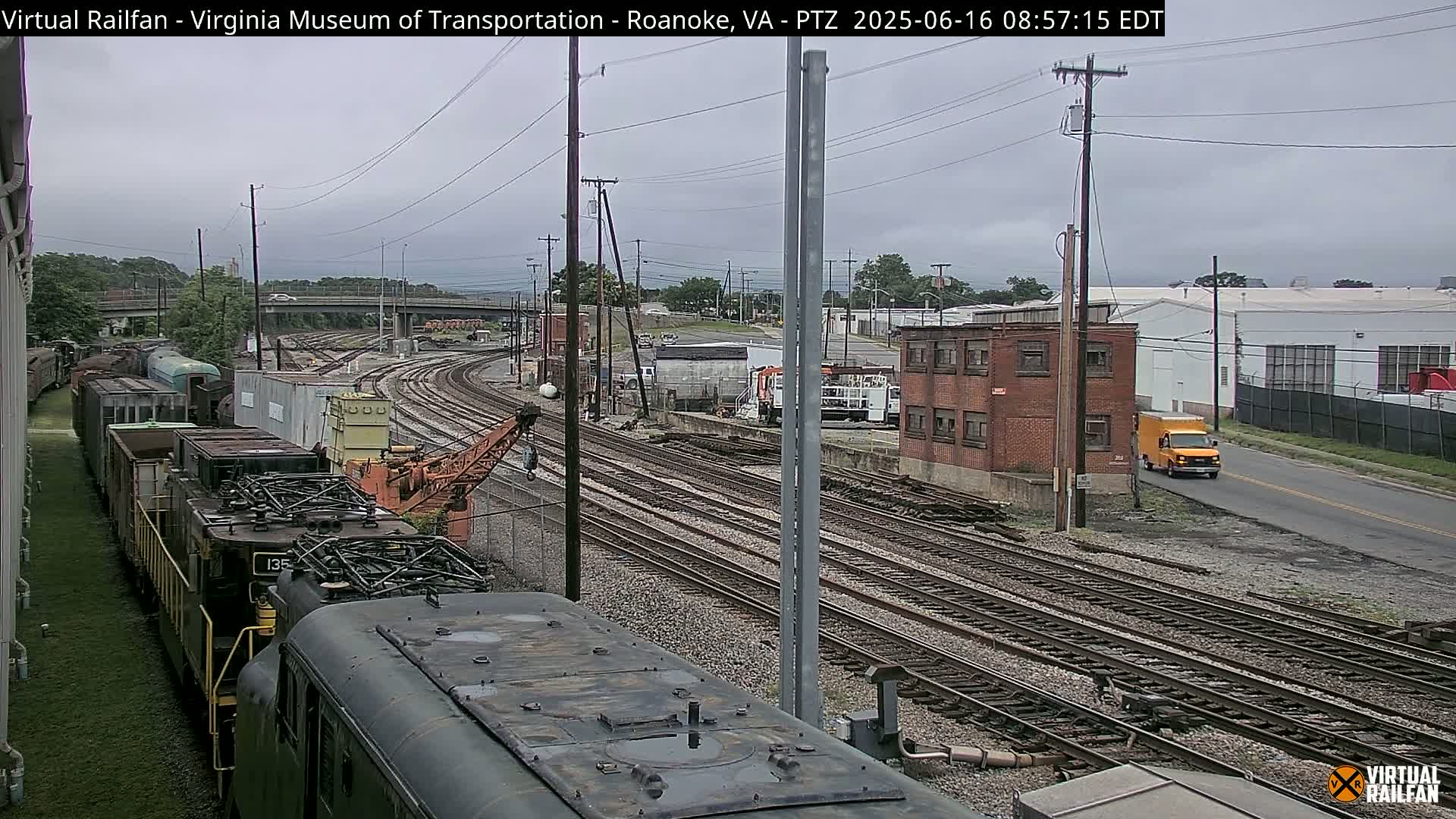 A freight train is parked next to a railway yard under an overcast sky, with multiple sets of railway tracks and industrial buildings visible in the background.